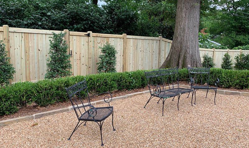 Black metal patio furniture on a gravel patio, in front of a hedge and wooden fence near a large tree.