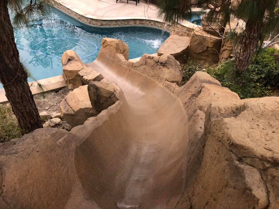A water slide is going down a rocky hill next to a swimming pool.