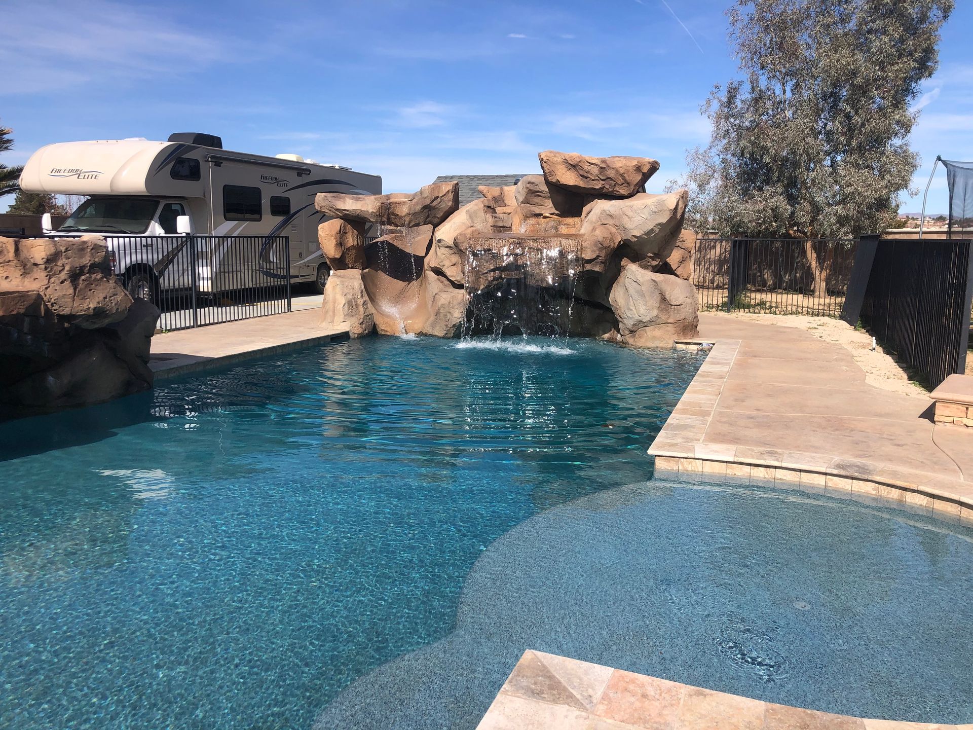A large swimming pool with a waterfall and a rv parked in the background.