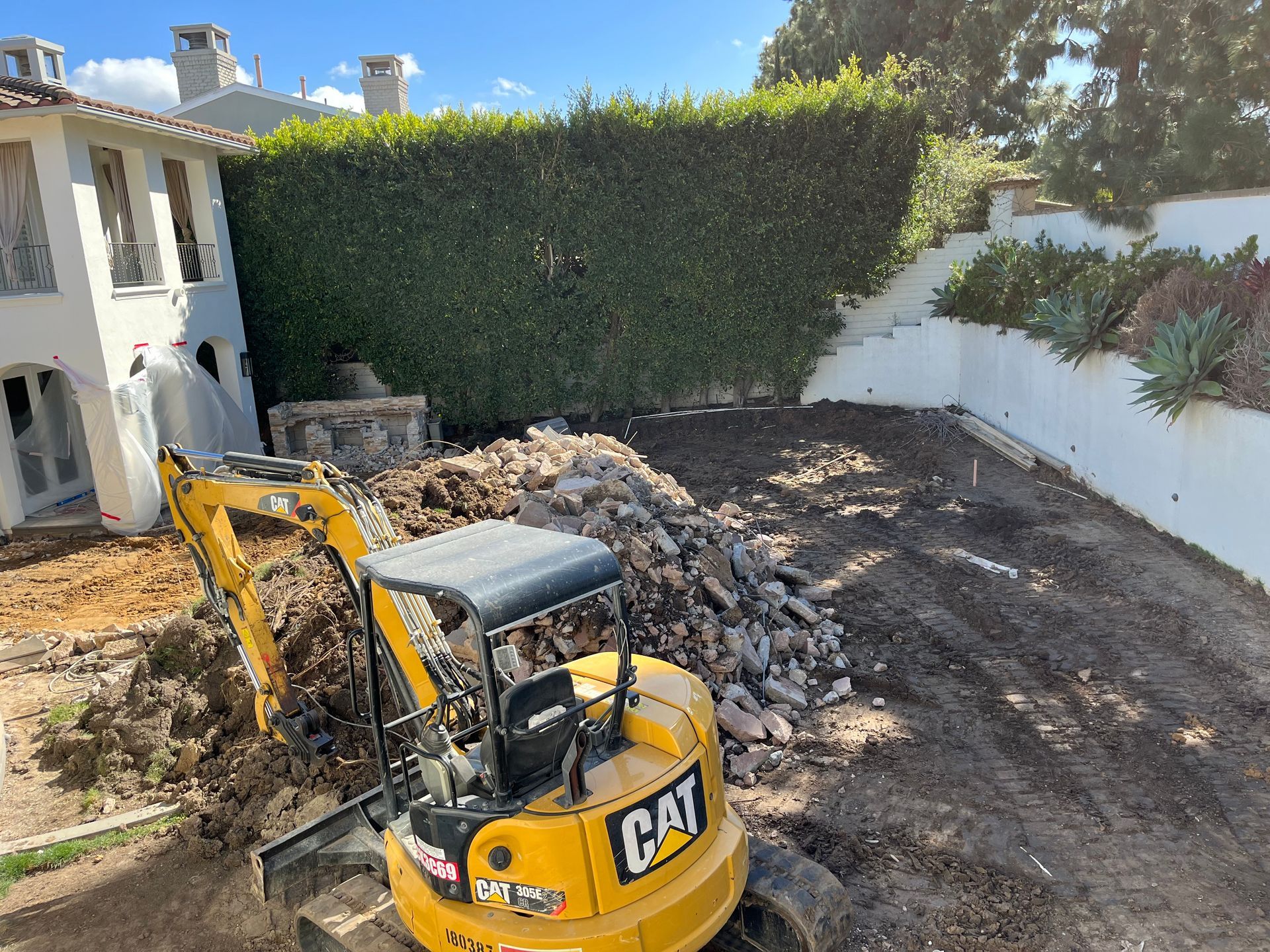 A yellow cat excavator is digging a hole in the dirt in front of a house.