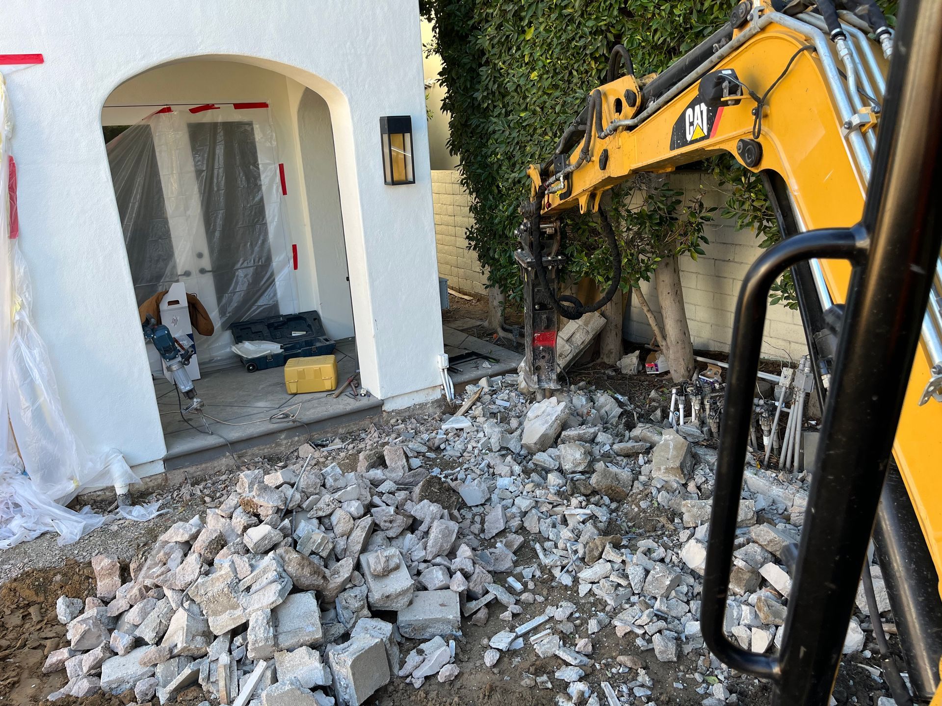 A yellow excavator is breaking concrete in front of a white house.