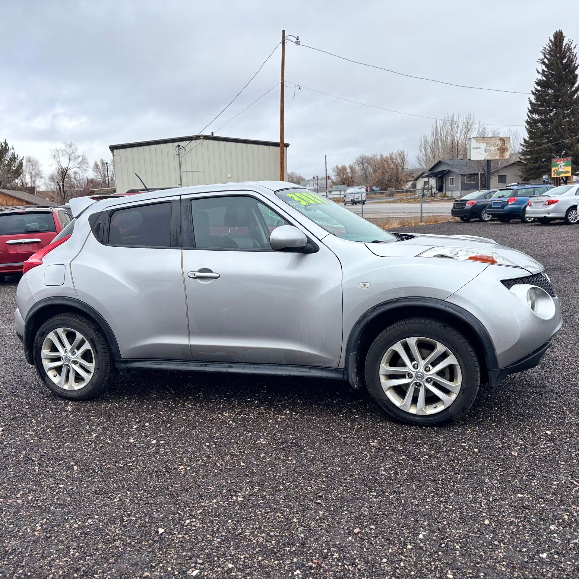 Silver Nissan Juke parked on gravel in a car lot on a cloudy day.