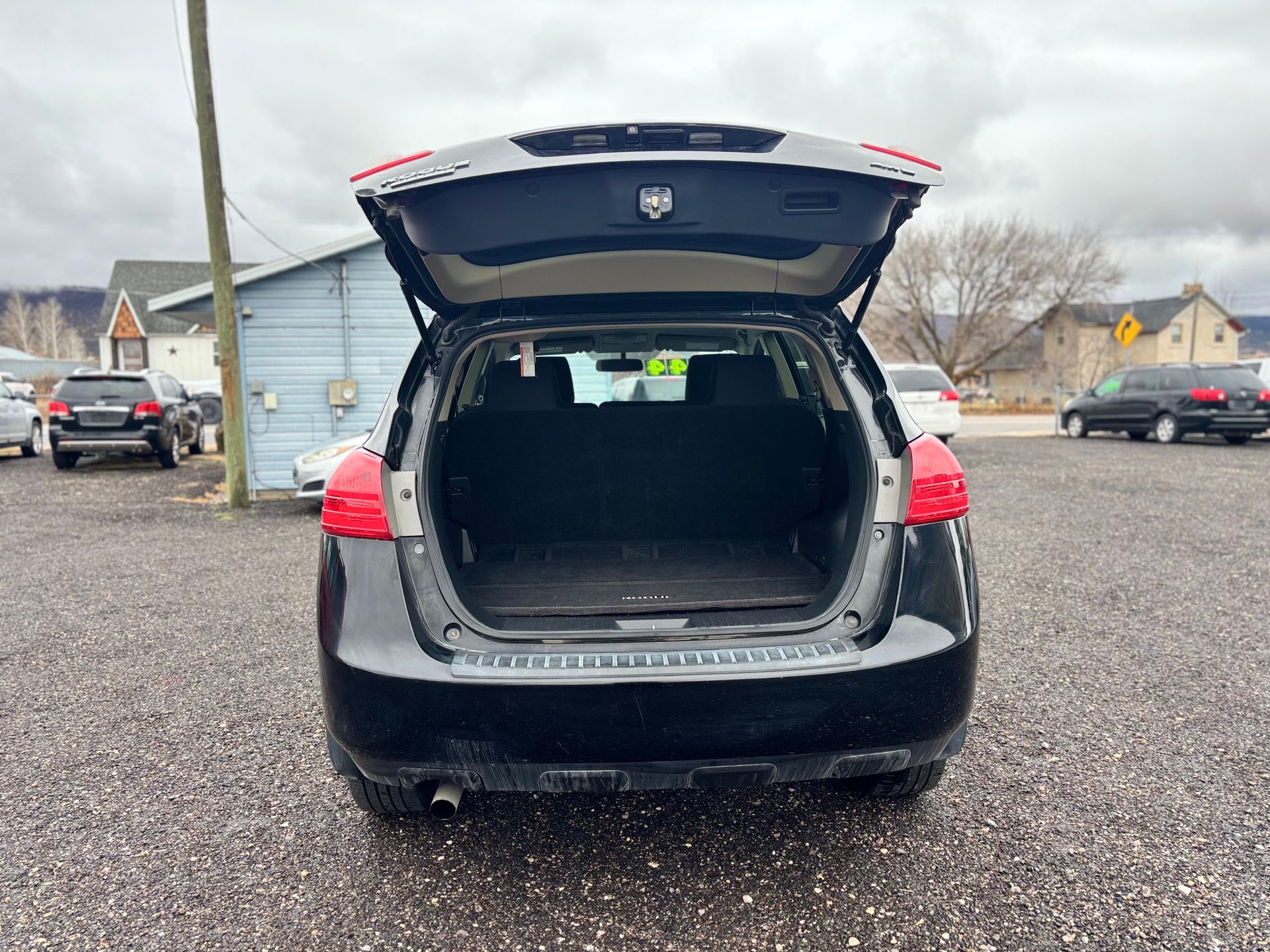 Black SUV with open trunk, parked on gravel lot. Buildings and other cars in background.
