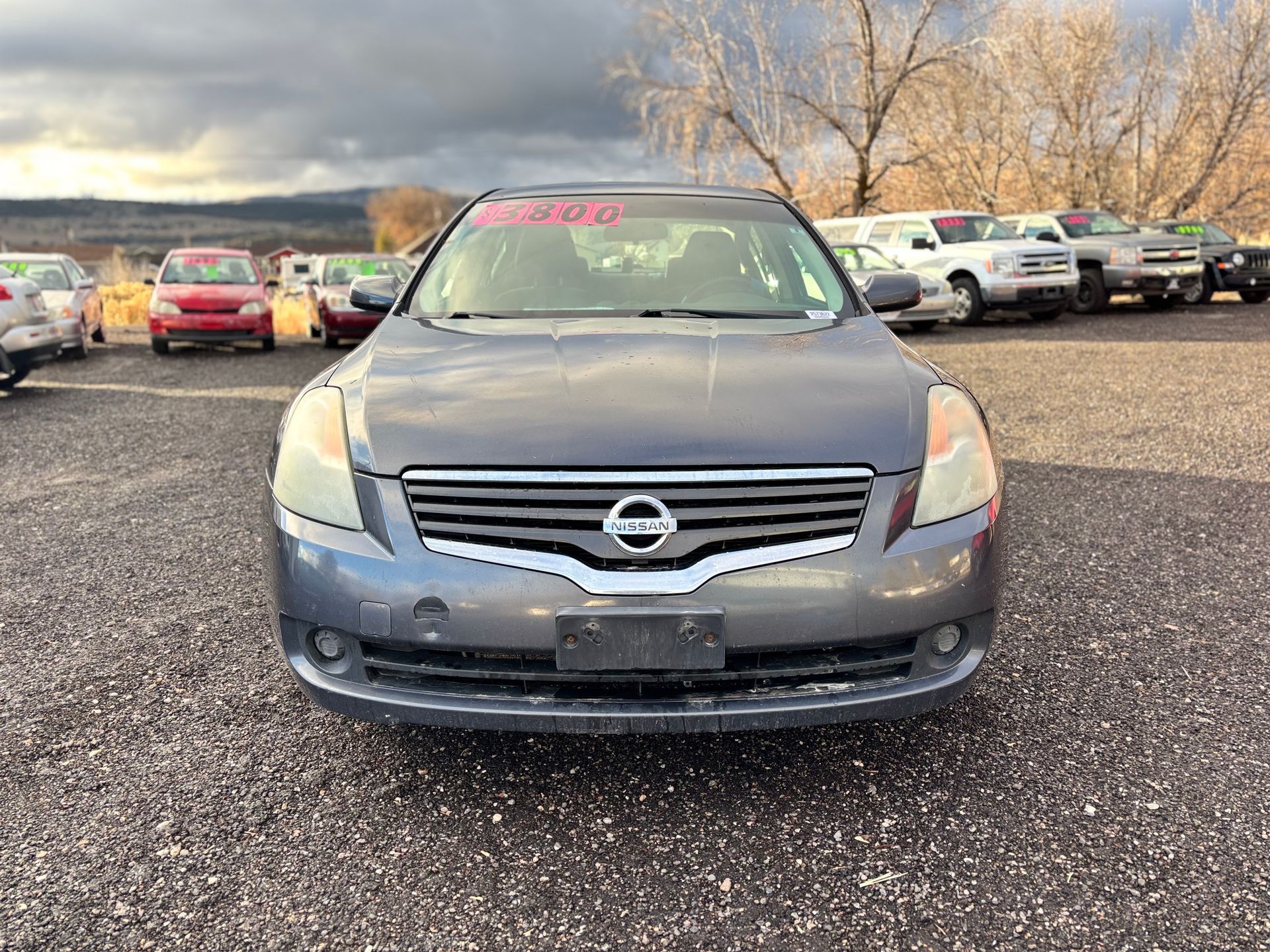 Gray Nissan Altima car on a gravel lot at a car dealership under a cloudy sky.