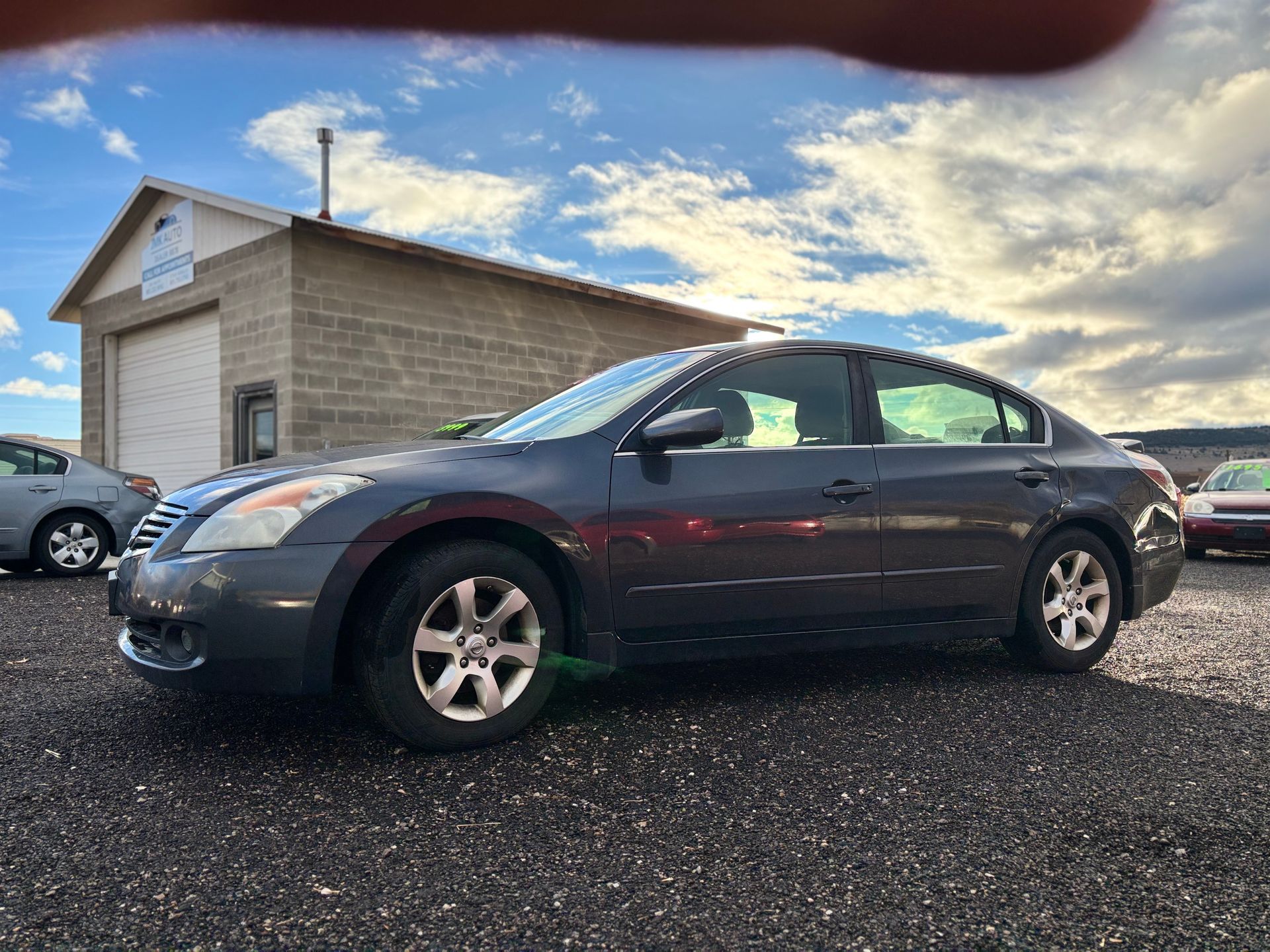 Gray Nissan Altima parked on gravel lot, building in background. Cloudy sky overhead.