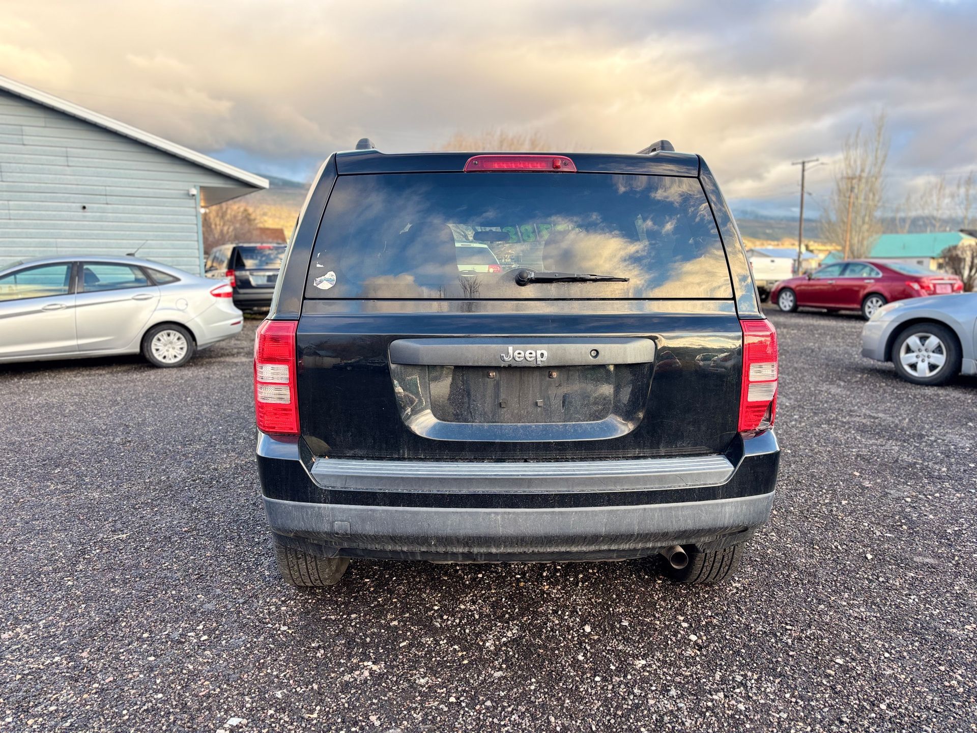 Black SUV, rear view, parked on gravel lot with other cars, cloudy sky in background.