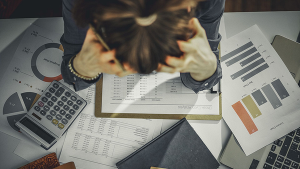 Top view of a stressed business person working at a cluttered desk filled with papers, a calculator, a notebook, and charts.