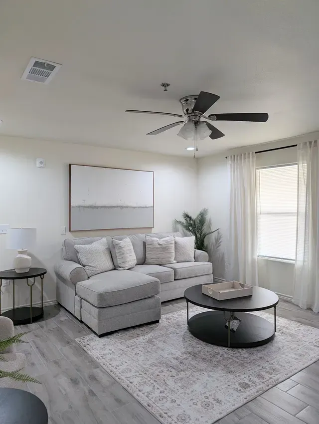 Bright living room with gray sofa, round coffee table, patterned rug, wall art, and ceiling fan near a window.
