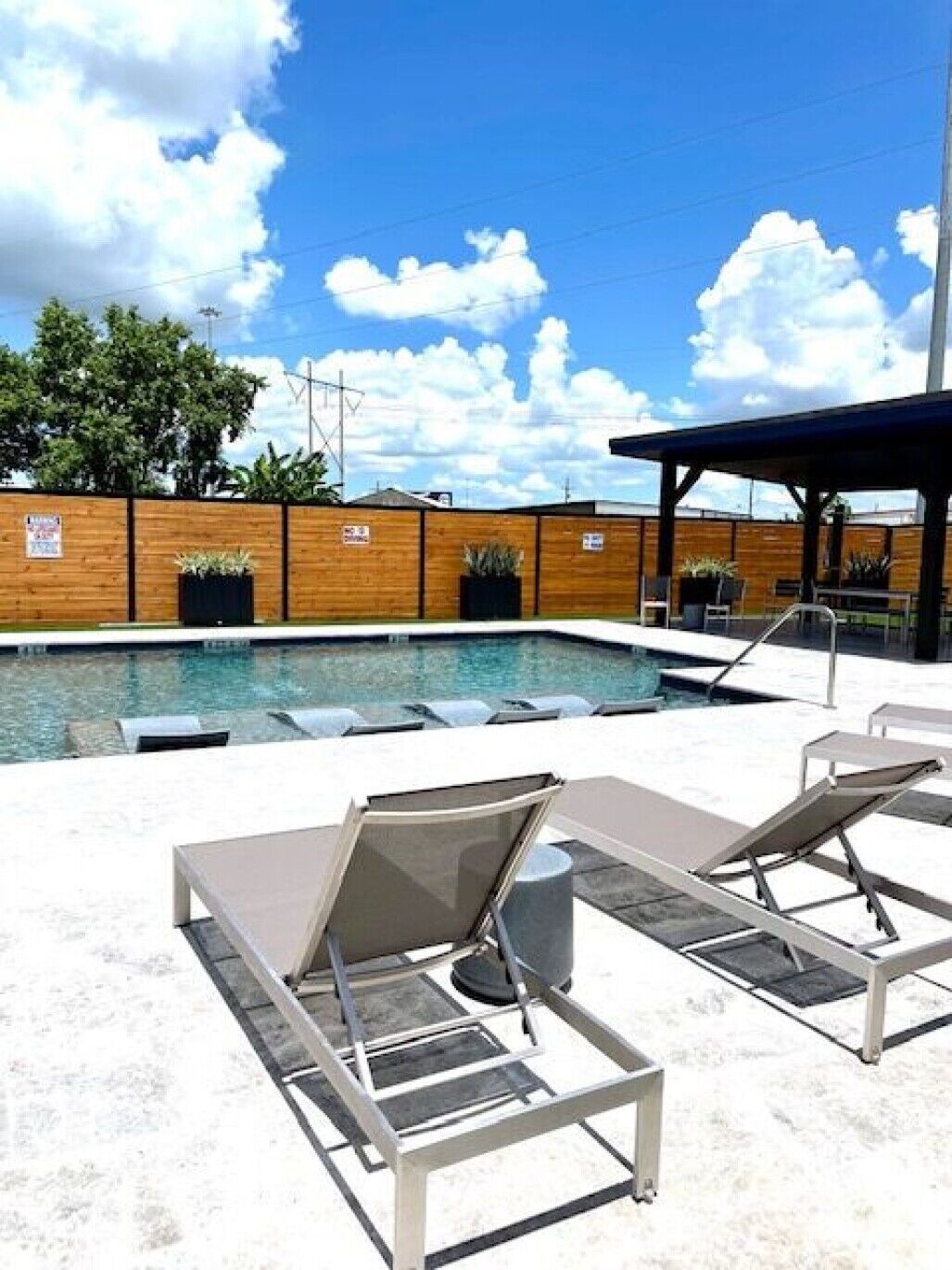 Rooftop pool with lounge chairs, blue sky, and potted trees behind a wooden fence.