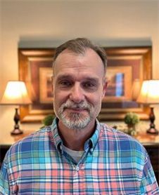 A man with a beard is wearing a plaid shirt and standing in front of a picture.