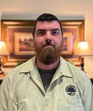 A man with a beard is wearing a plaid shirt and standing in front of a picture.