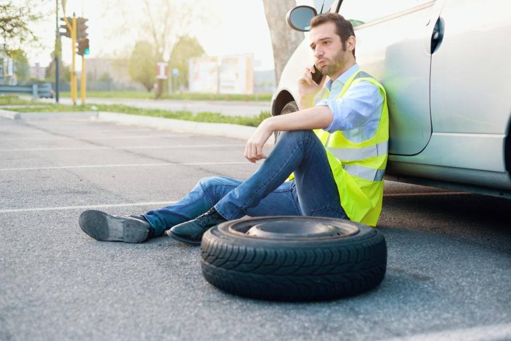 A Man Is Sitting on The Ground Next to A Tire and Talking on A Cell Phone — Alfie's Towing In Bentley Park, QLD