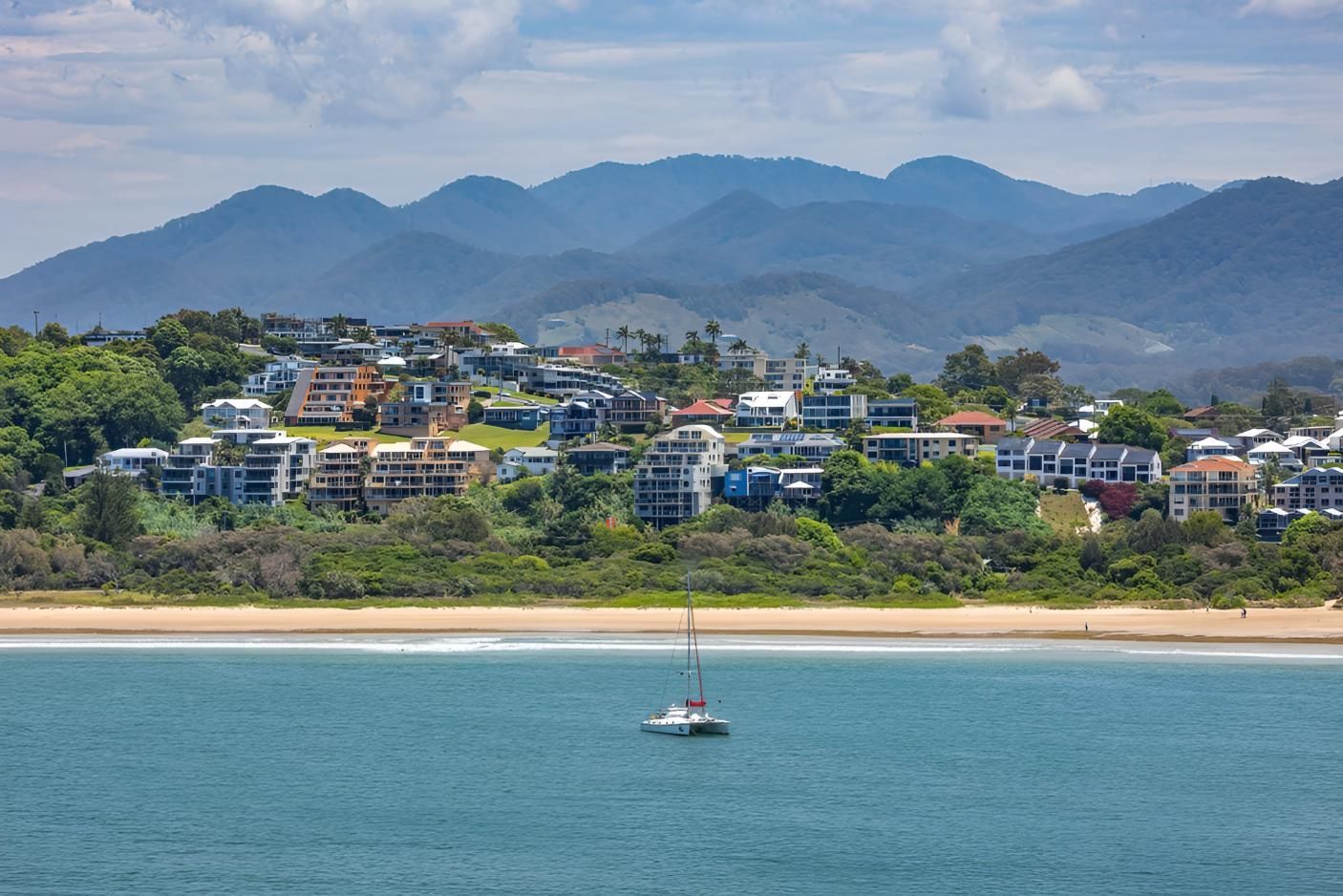 A Sailboat is Floating on Top of a Body of Water With Mountains in the Background — Coastal Test & Tag in Coffs Harbour, NSW