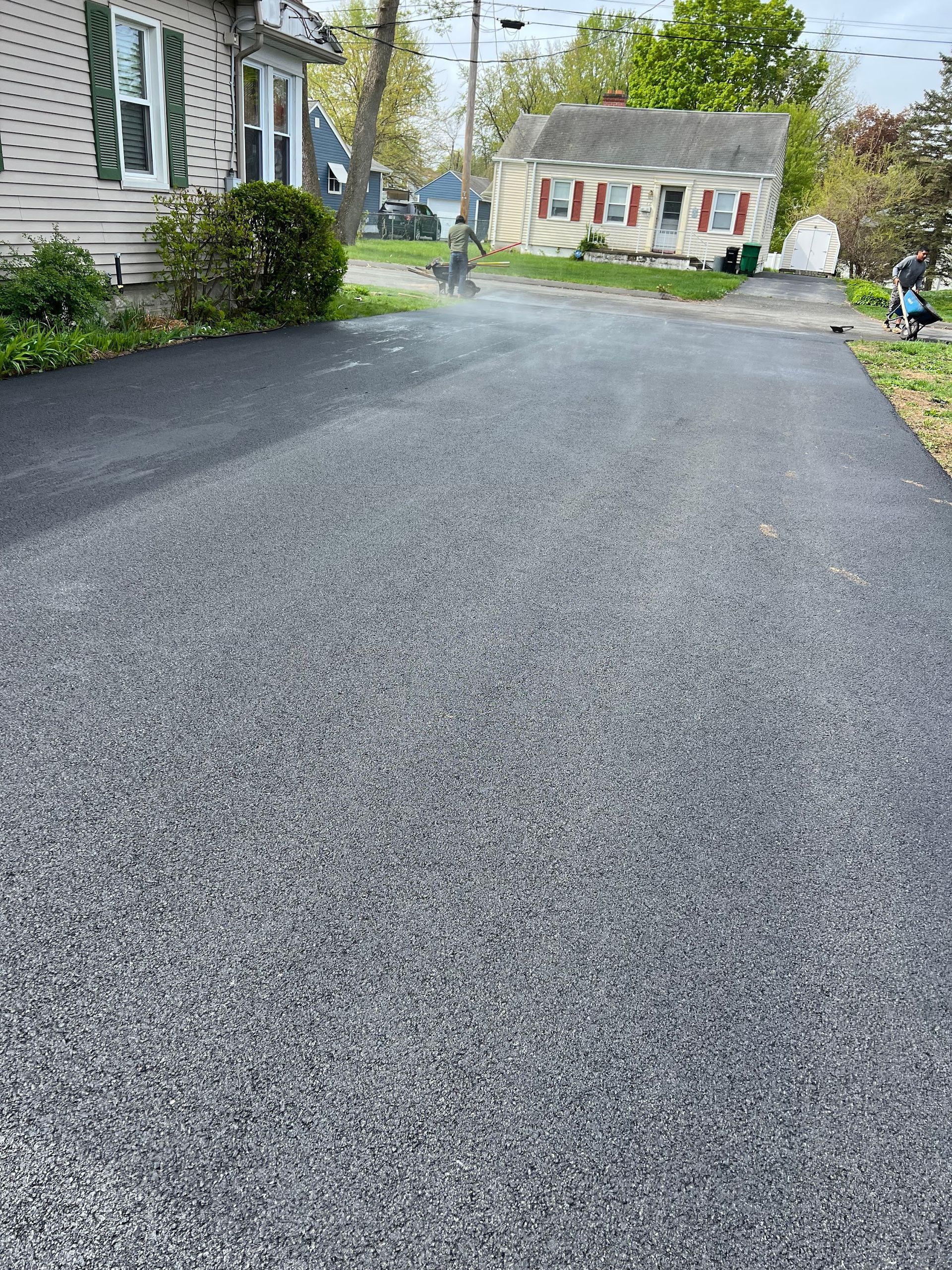 A asphalt driveway leading to a house with a white house in the background.