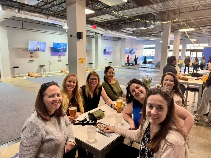 Group of people smiling at a table in a bright warehouse-style café, with drinks and snacks.