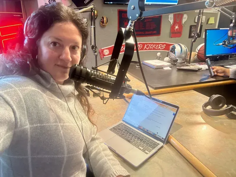 Woman speaking into a microphone at a desk with a laptop in a radio studio.