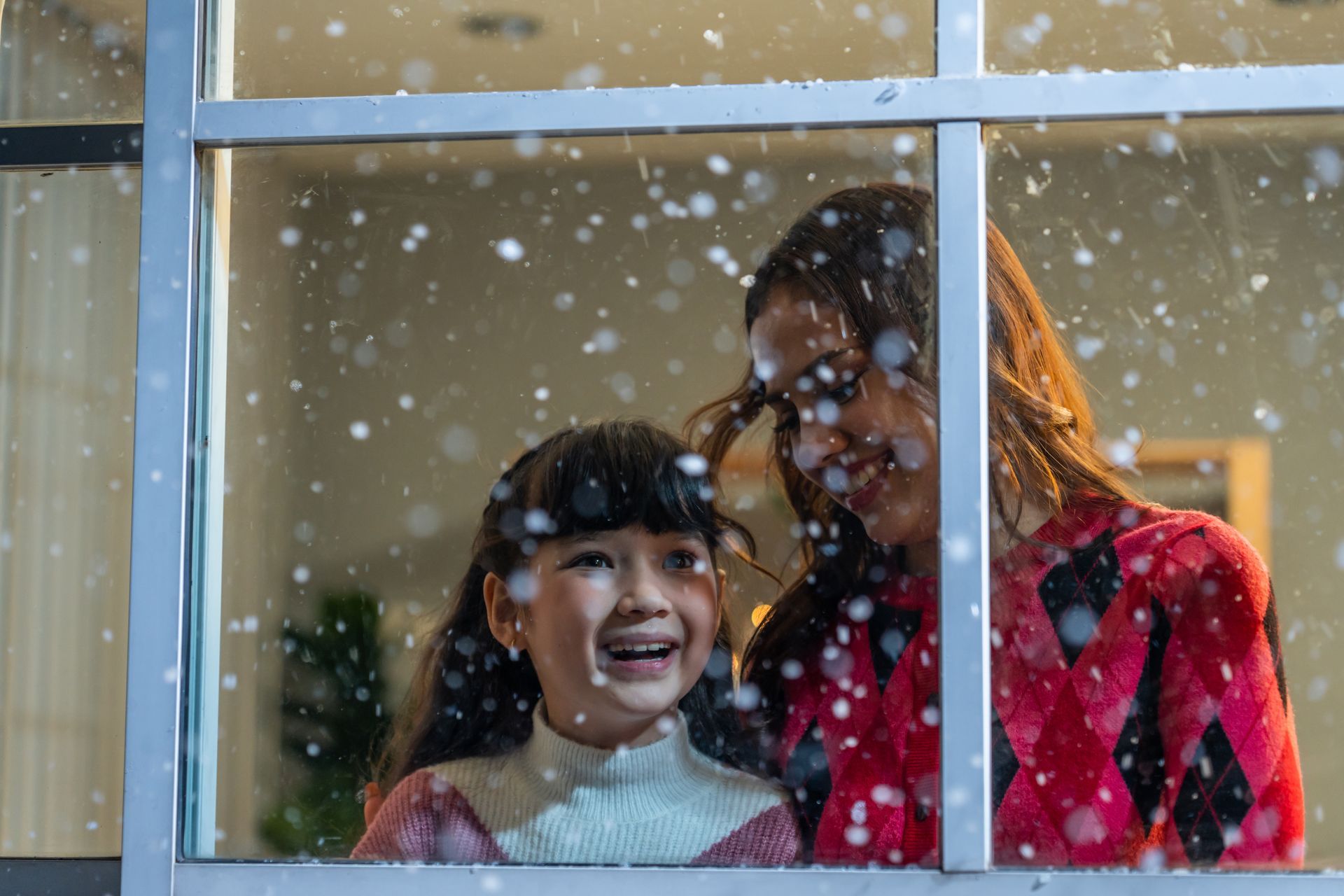 Woman and child looking through window at snowfall, smiling.