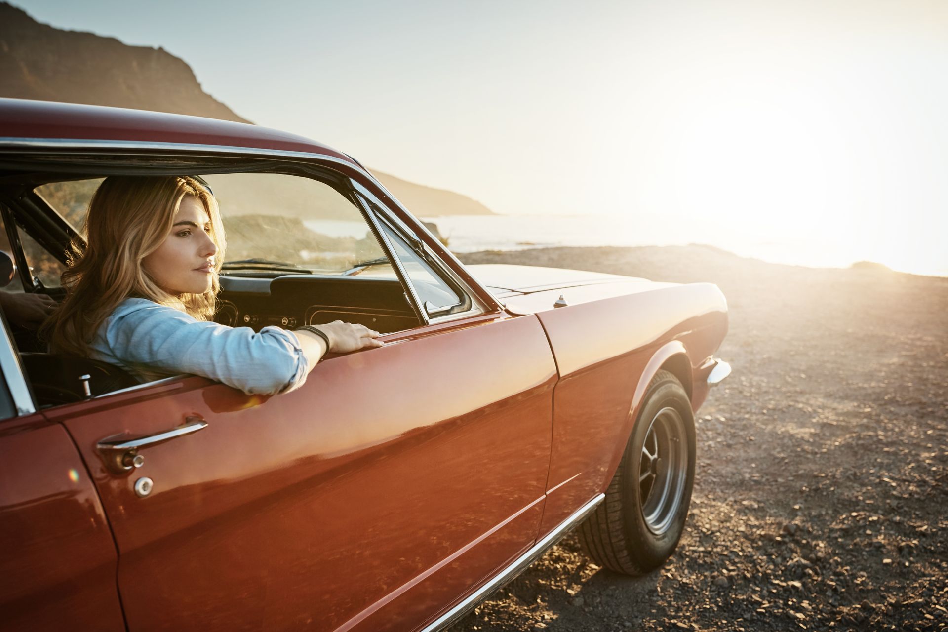 Woman in a red classic car looking out at a coastal view; sunlight.