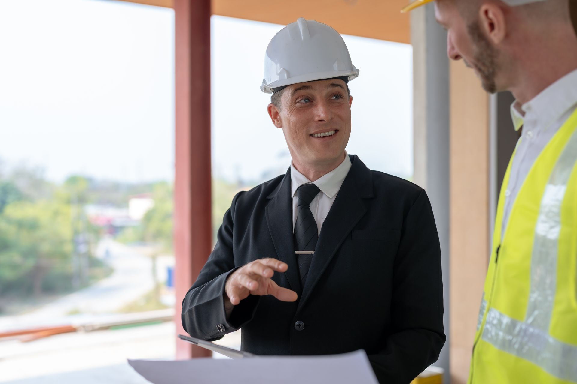 Two construction workers in hard hats, one in a suit, discussing blueprints at a construction site.