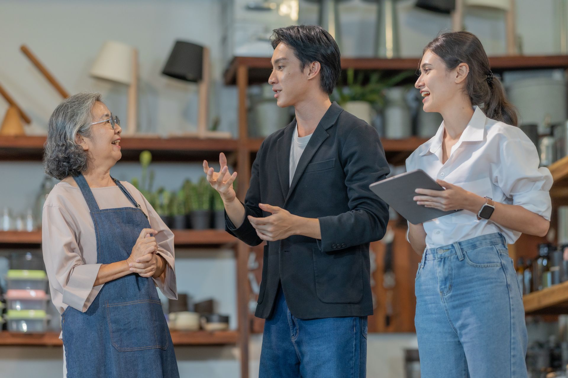 Three people in a shop, talking: a woman in an apron, a man in a blazer, and a woman with a tablet.