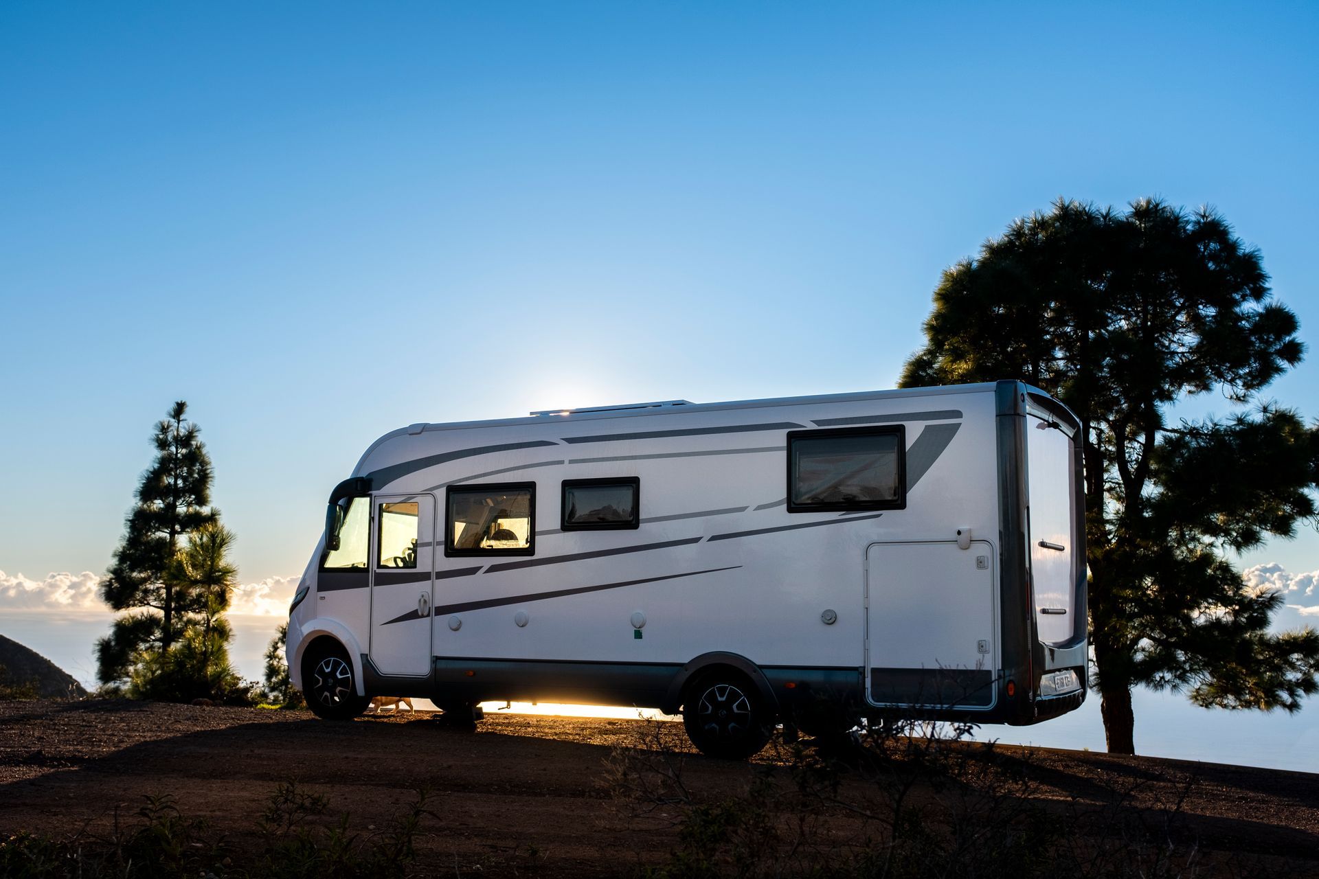 White RV parked on a hilltop with trees, sun setting in the background.