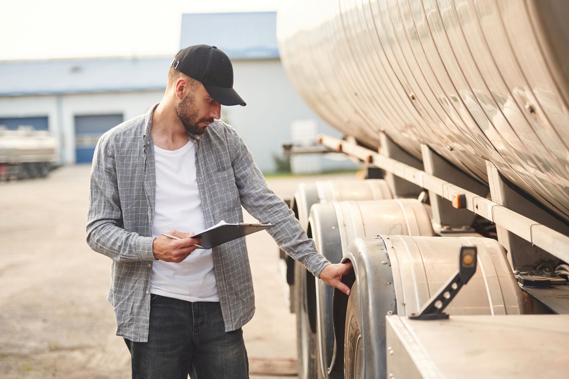 Trucker in black cap inspecting trailer tires, holding clipboard.
