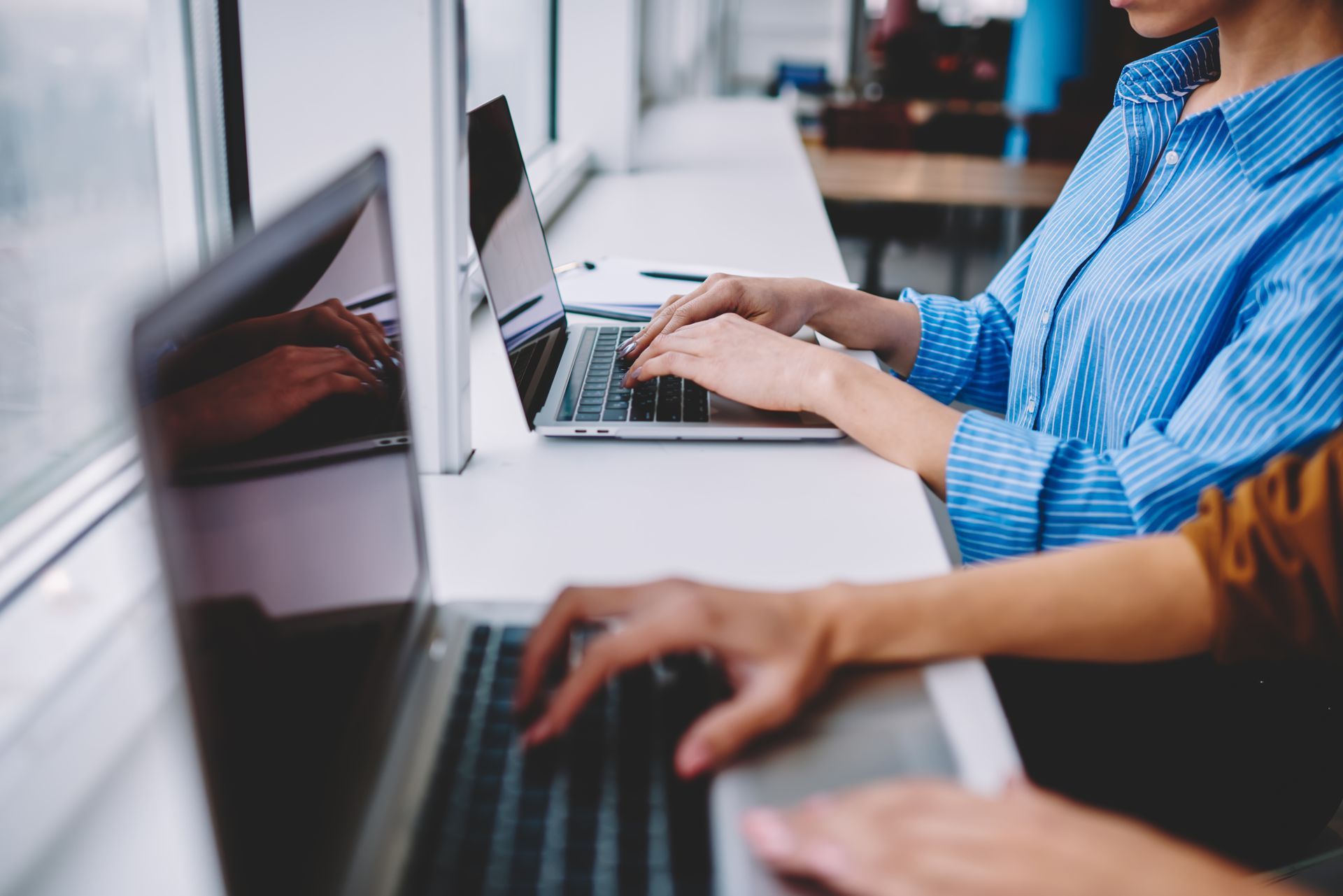 Two people typing on laptops at a white counter near a window. One person wears a blue shirt.