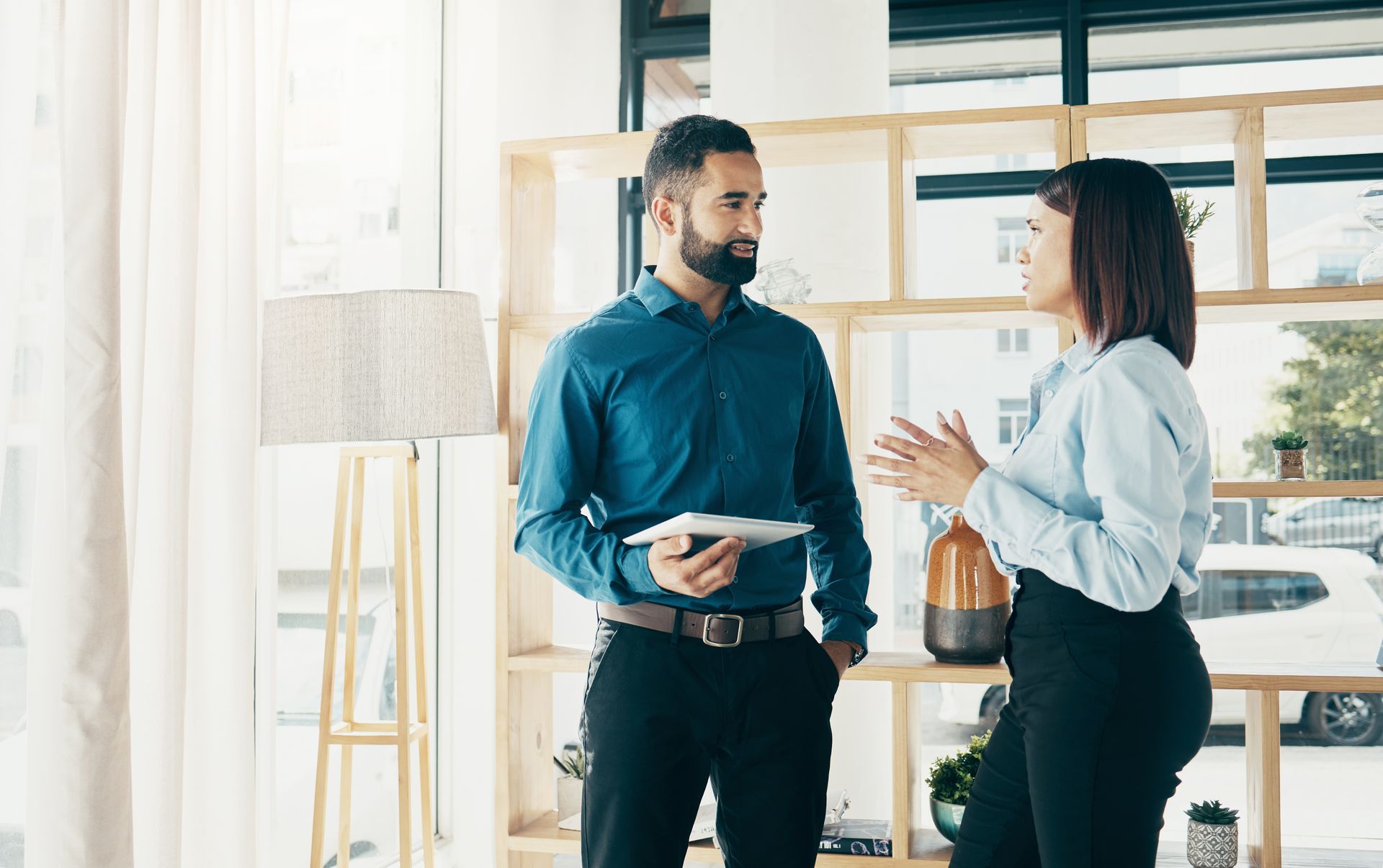Man and woman in office, conversing near window and shelf. Man holds tablet, woman gestures.
