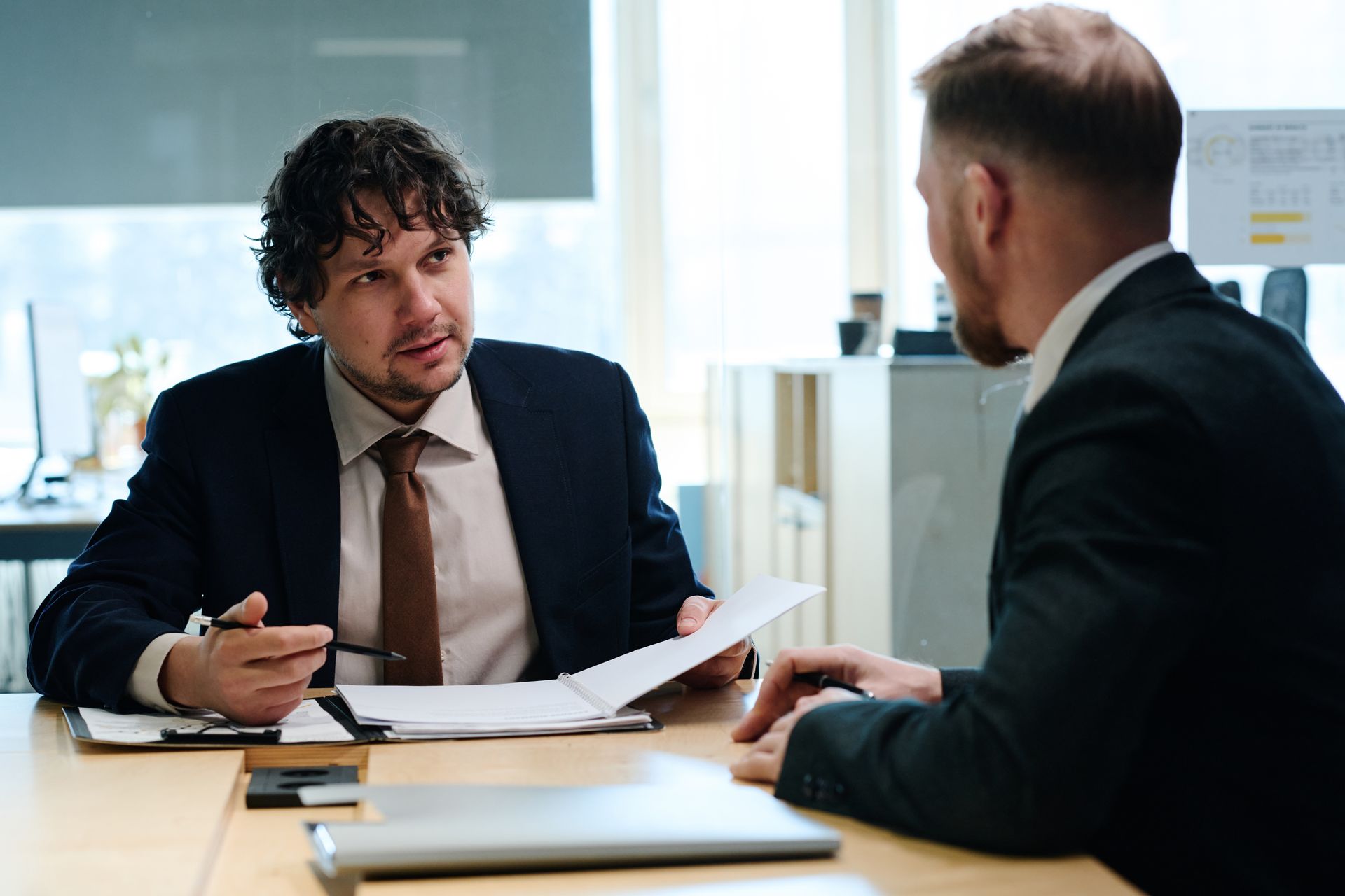 Two men in suits at a desk, reviewing documents. The man on the left points at the papers and speaks.