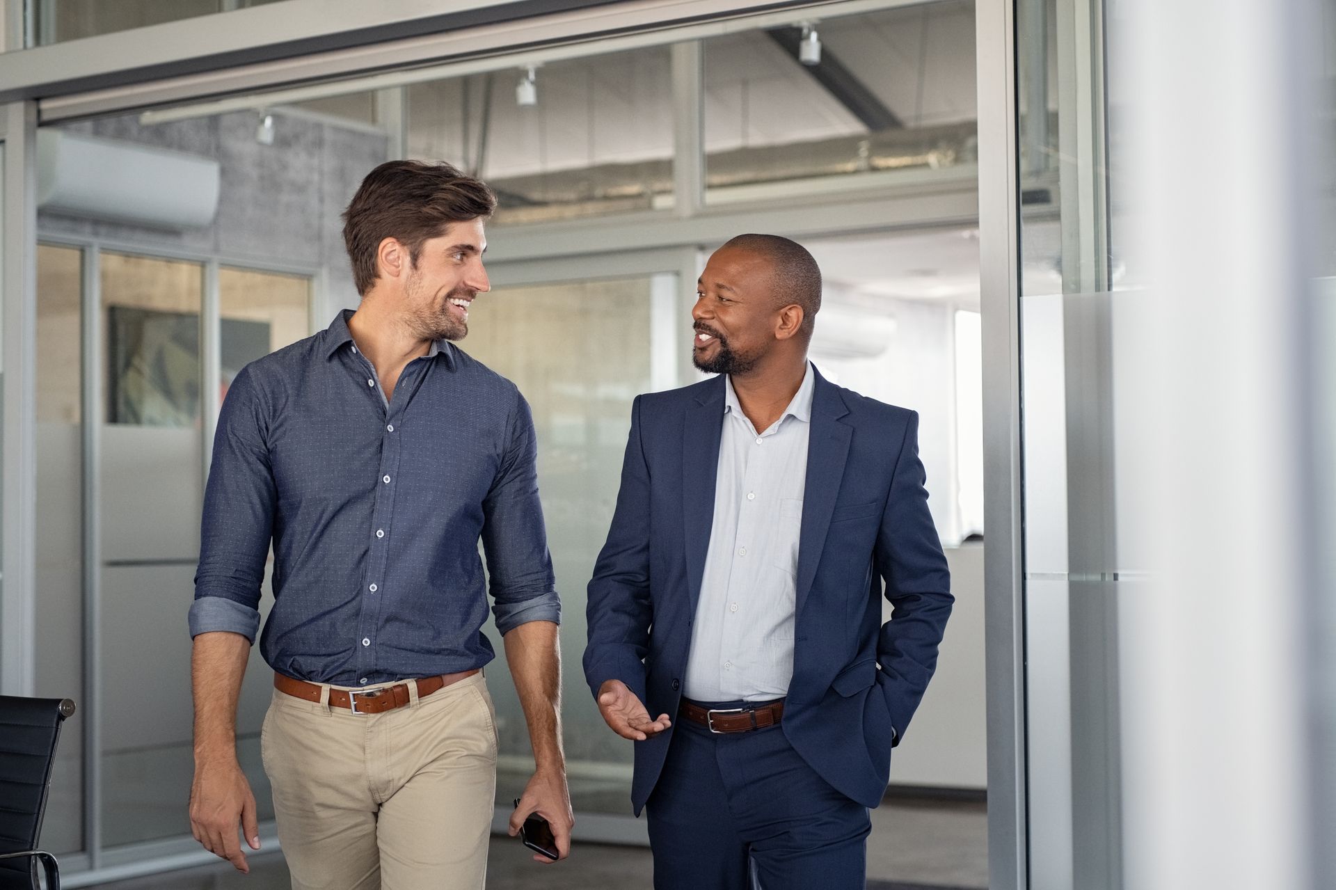 Two men in office attire walk and converse, smiling in a modern office.
