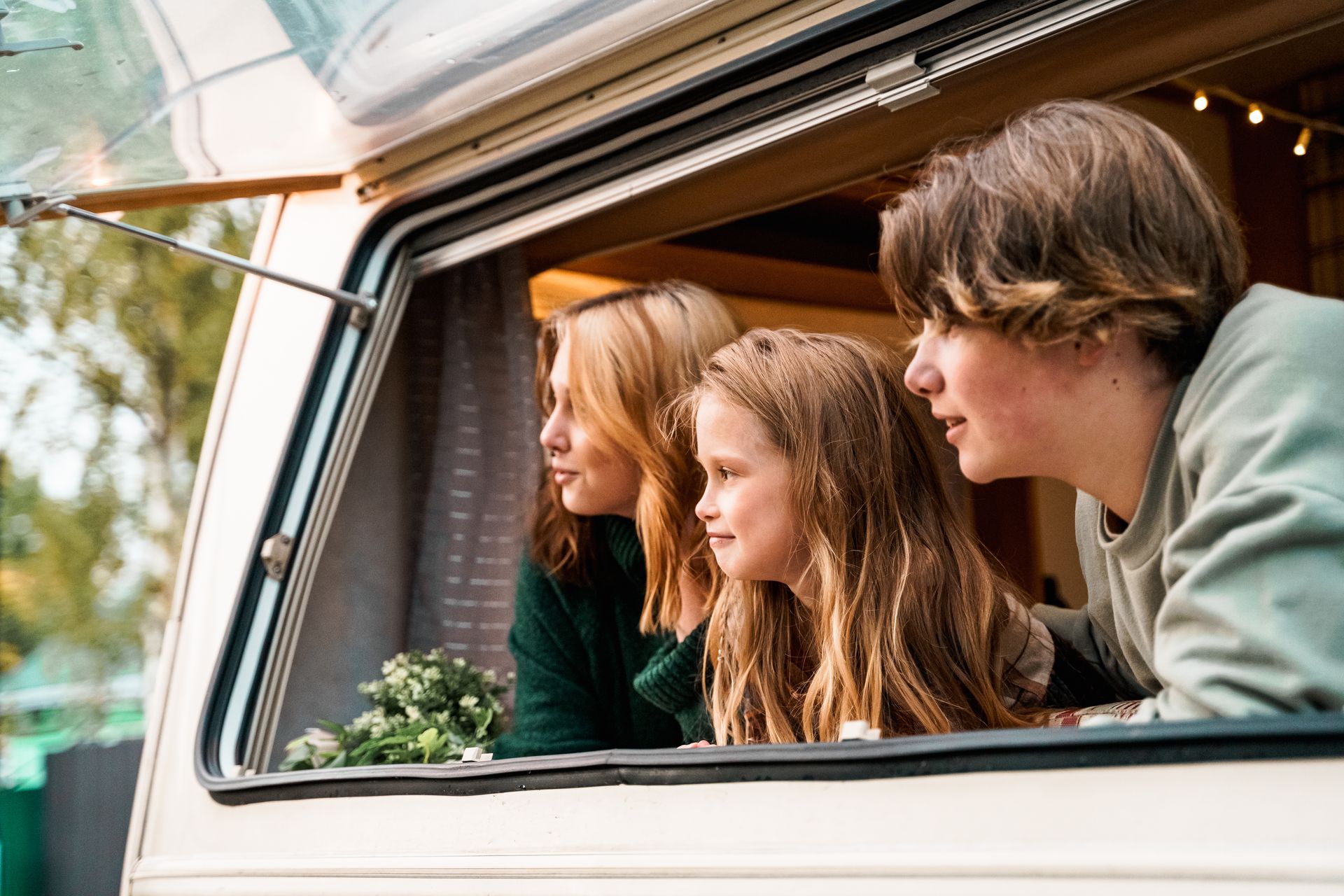 Three people looking out of a camper window: one with long blonde hair, one with long brown hair, and one with short brown hair.