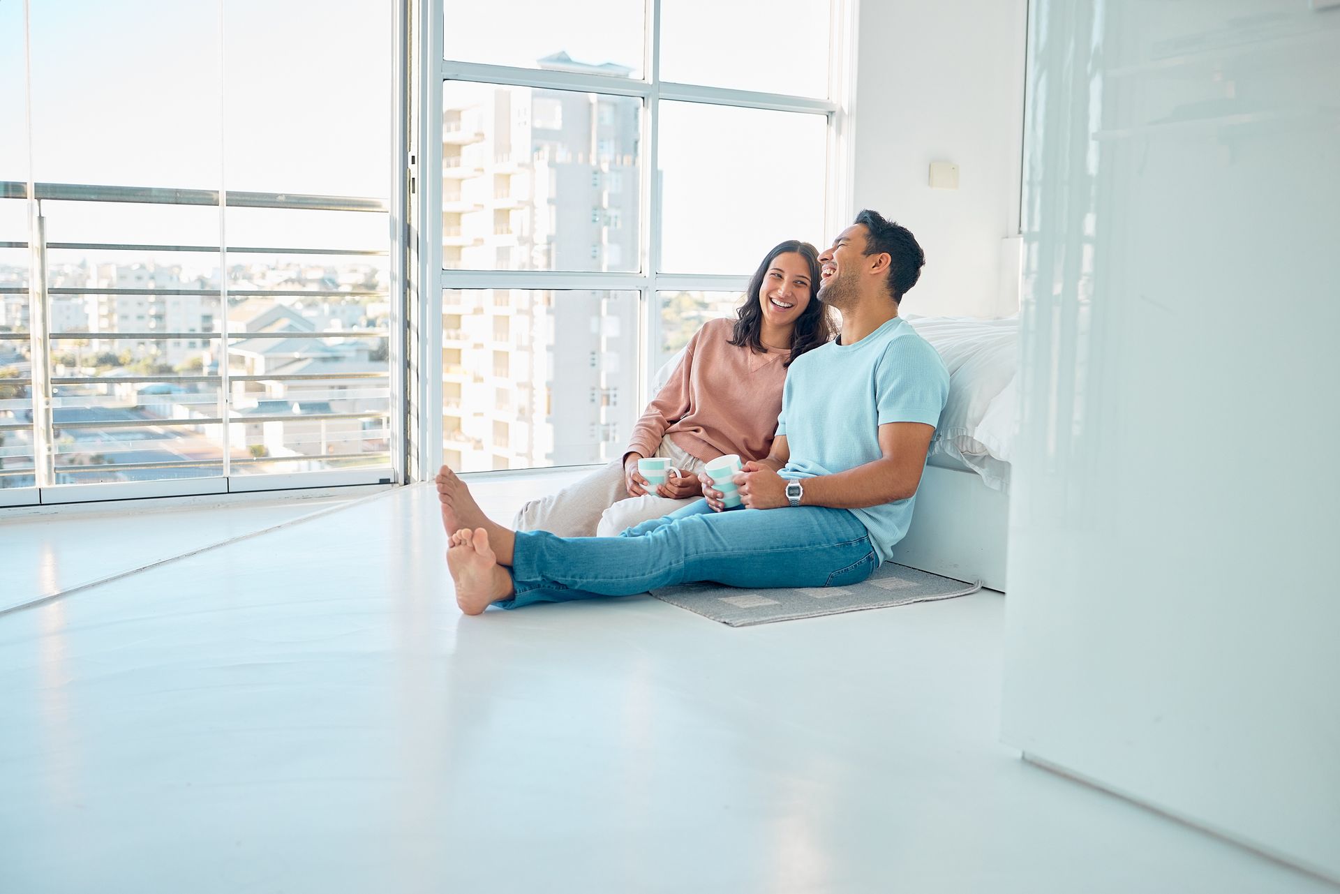 Couple smiling, sitting on floor by window, holding mugs. Bright, modern room with city view.