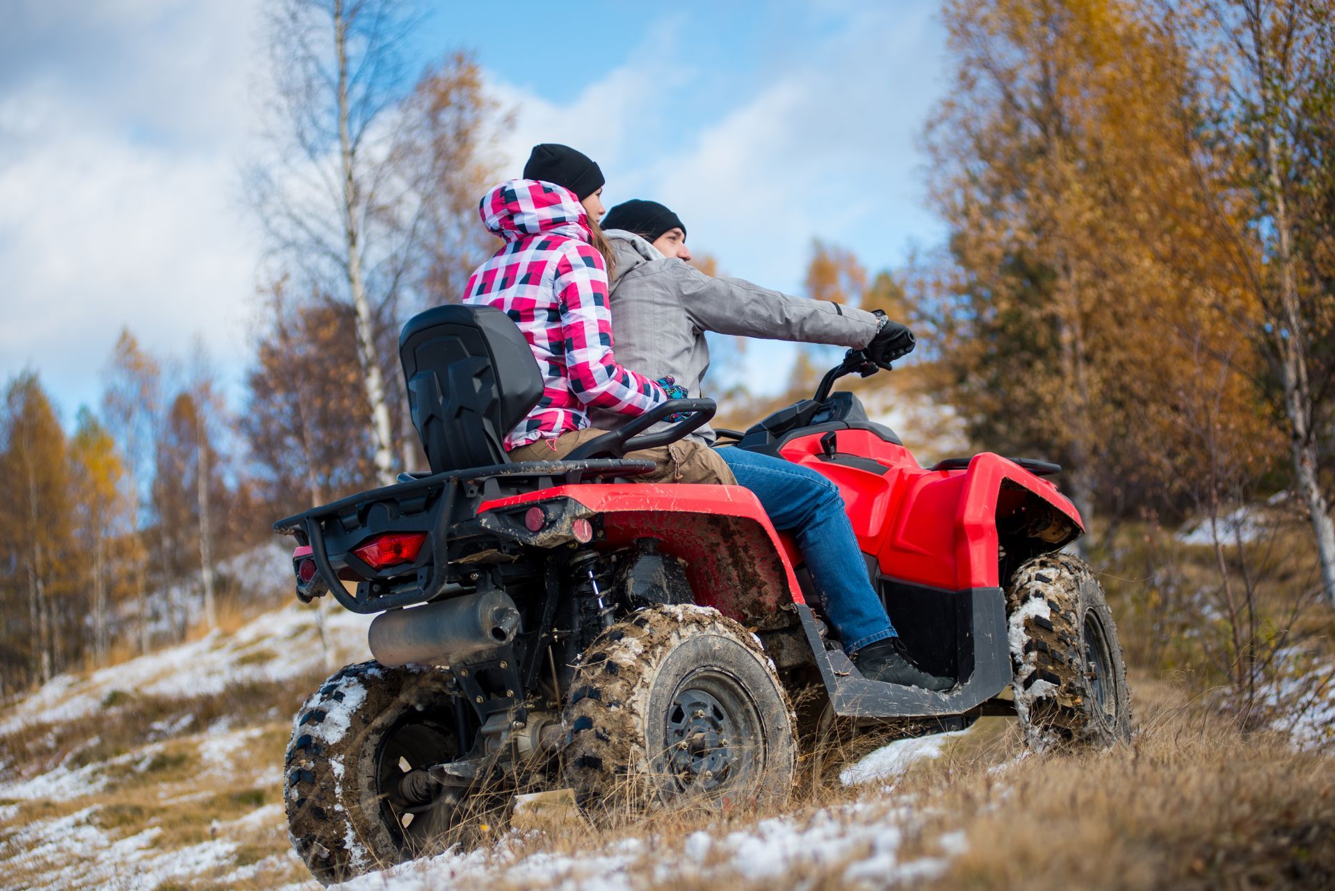 Couple riding a red ATV on a trail. They are wearing warm clothes, with trees in a fall setting.