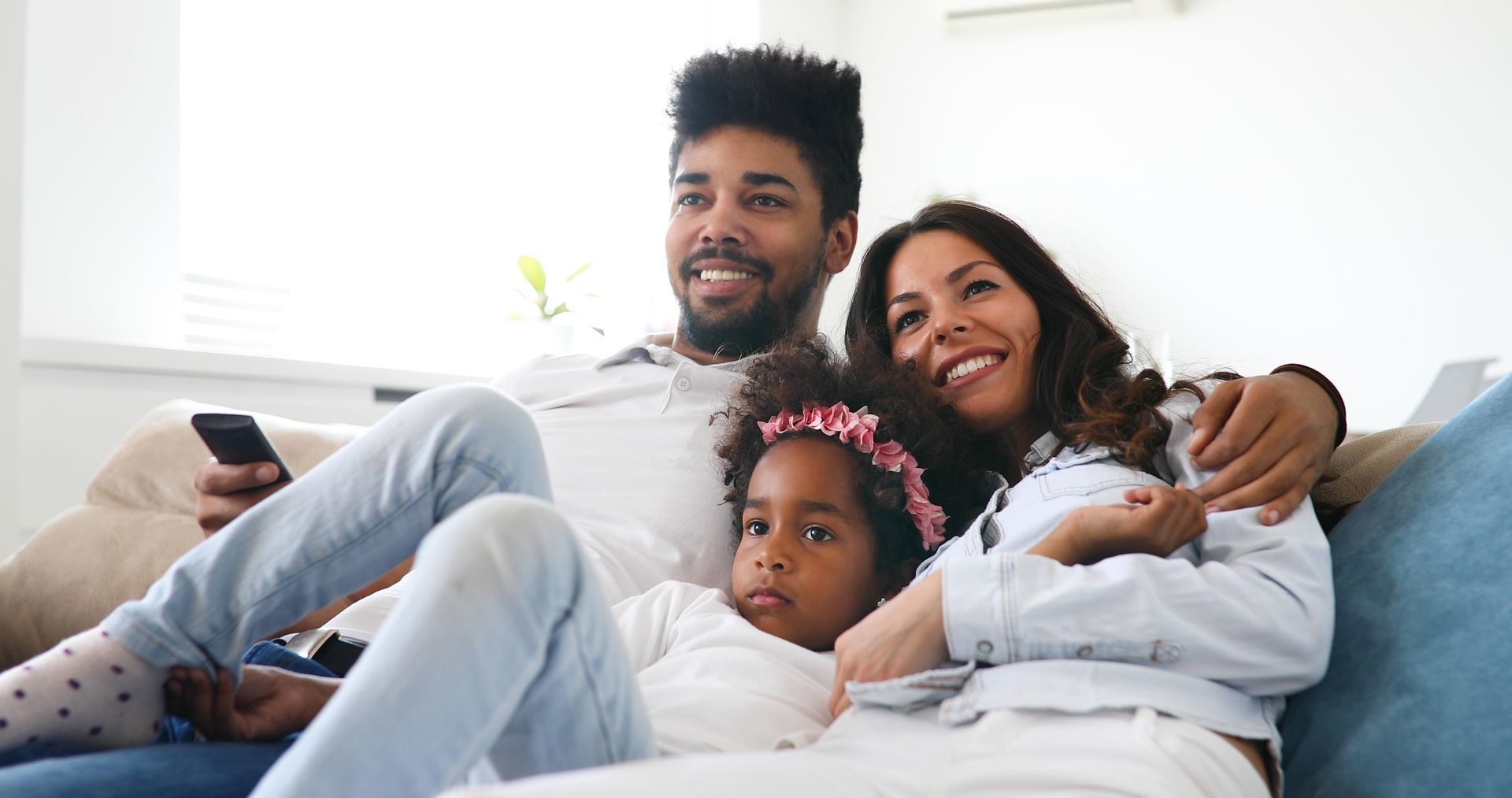 Family of three watching TV on a couch, smiling. Includes a child with a floral headband.