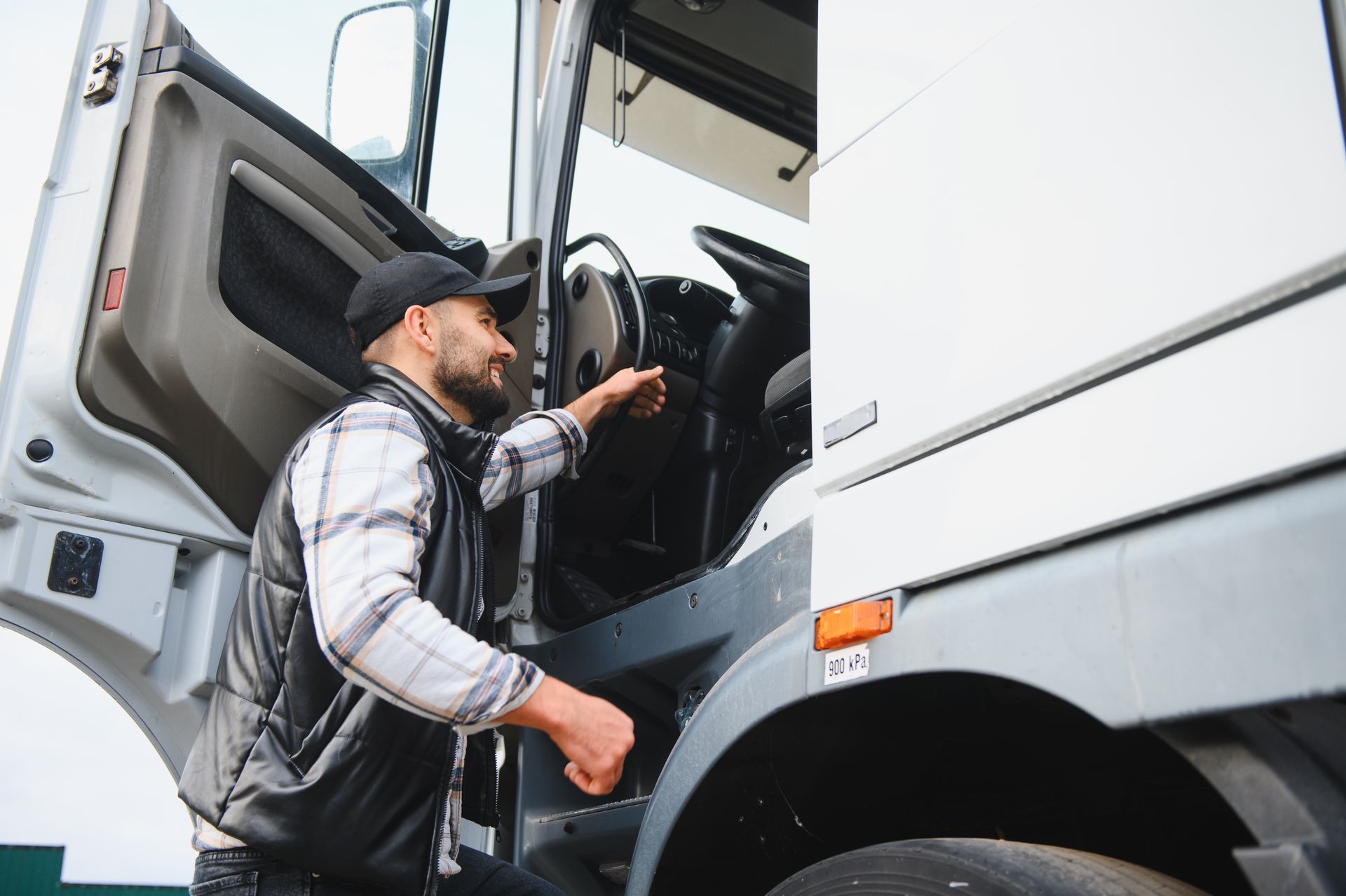 Truck driver entering the cab, smiling. Wearing cap, vest, and plaid shirt. Truck door open.
