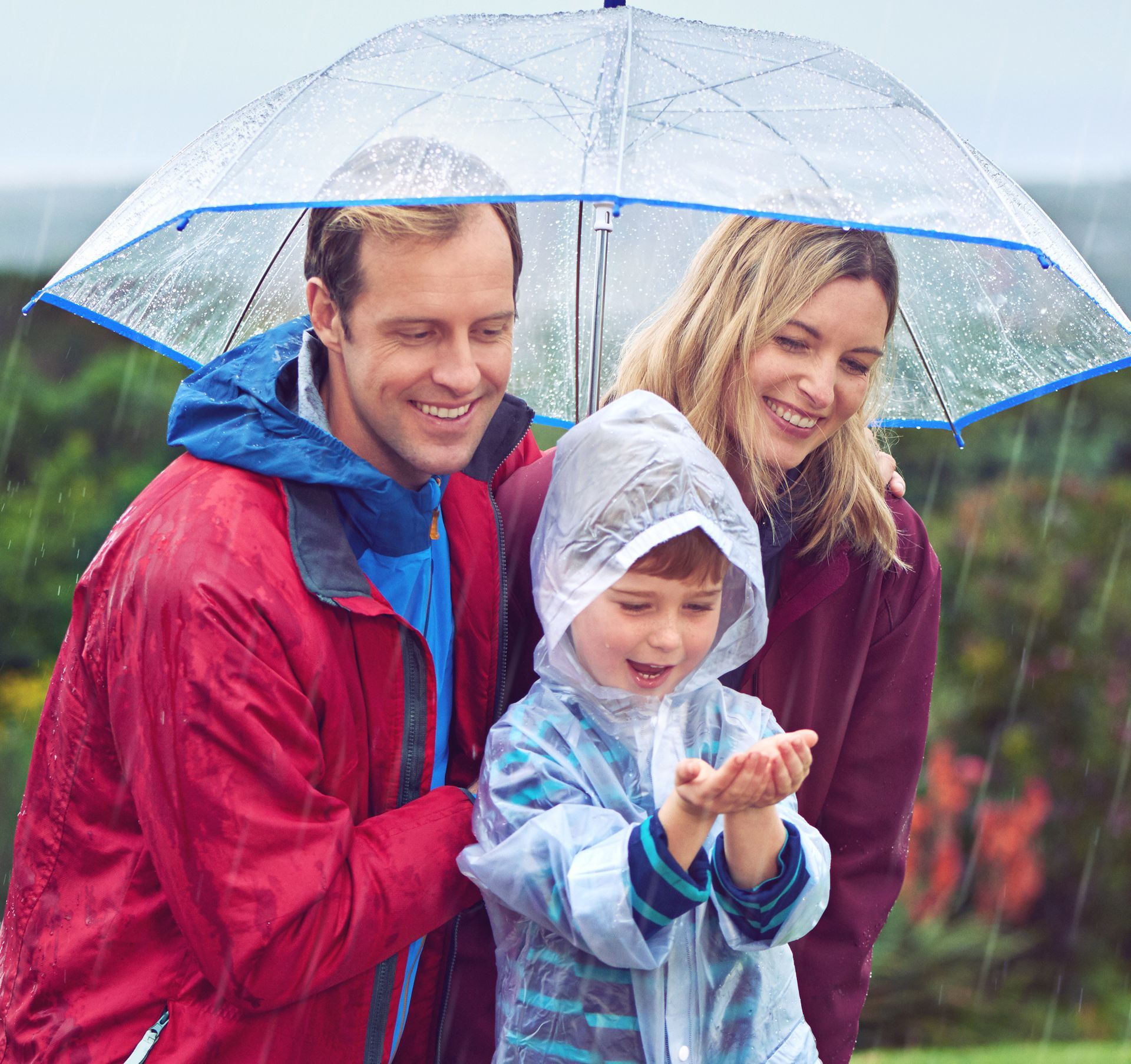 Family smiling under a clear umbrella in the rain; child in a raincoat catches raindrops.