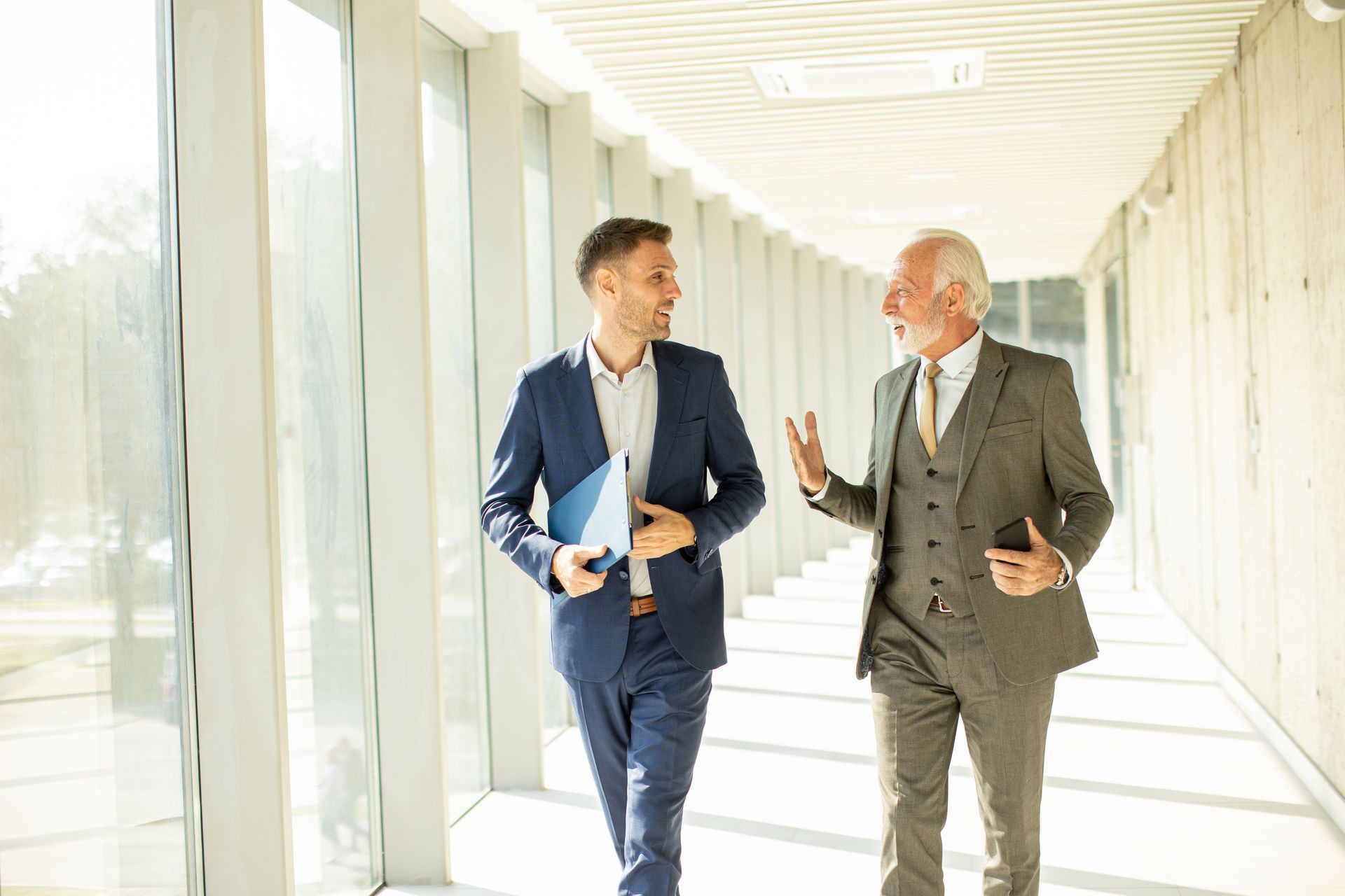Two men in suits walking and conversing in a modern hallway. The older man gestures while talking.