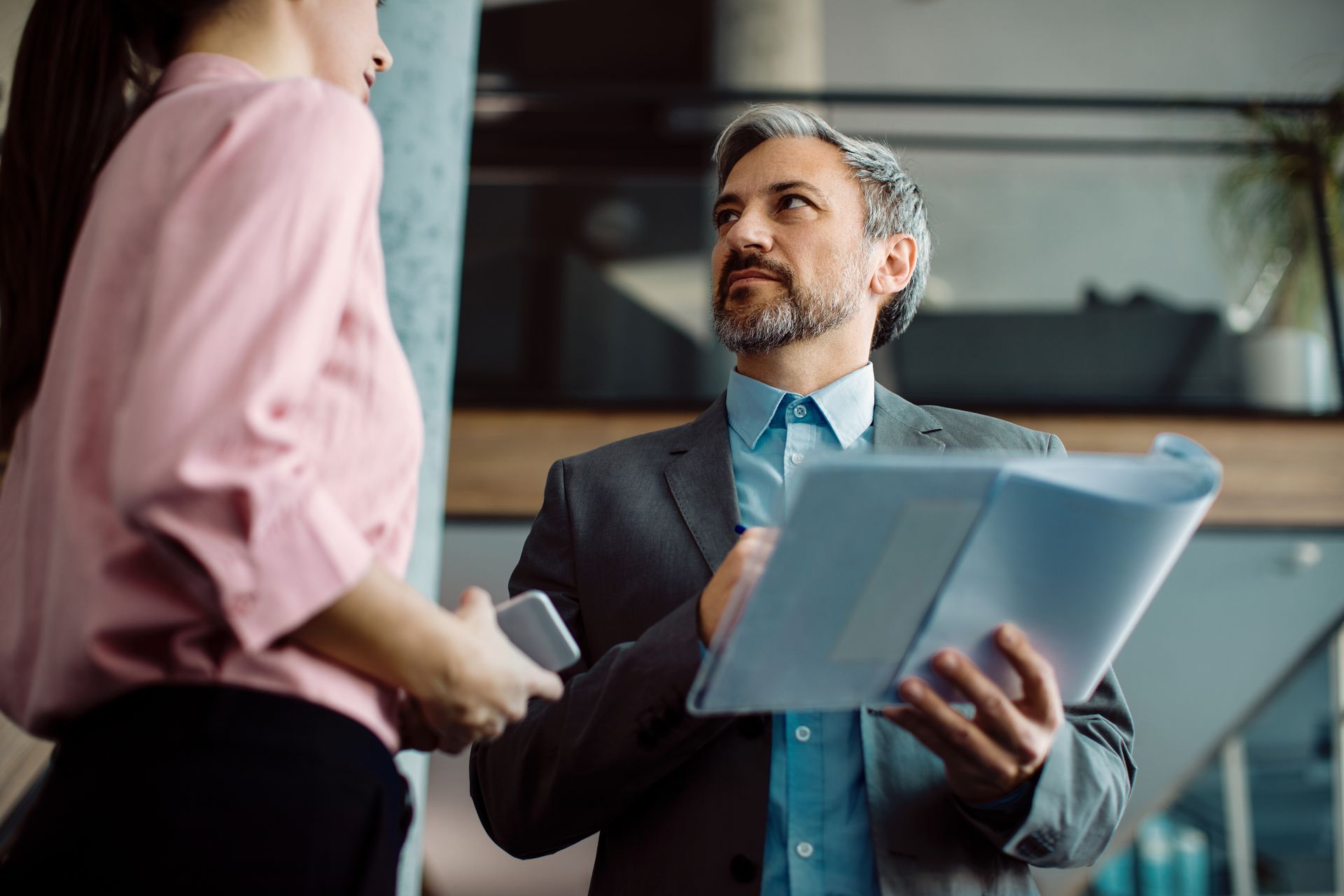 Man in suit discusses papers with woman in pink shirt, both indoors.