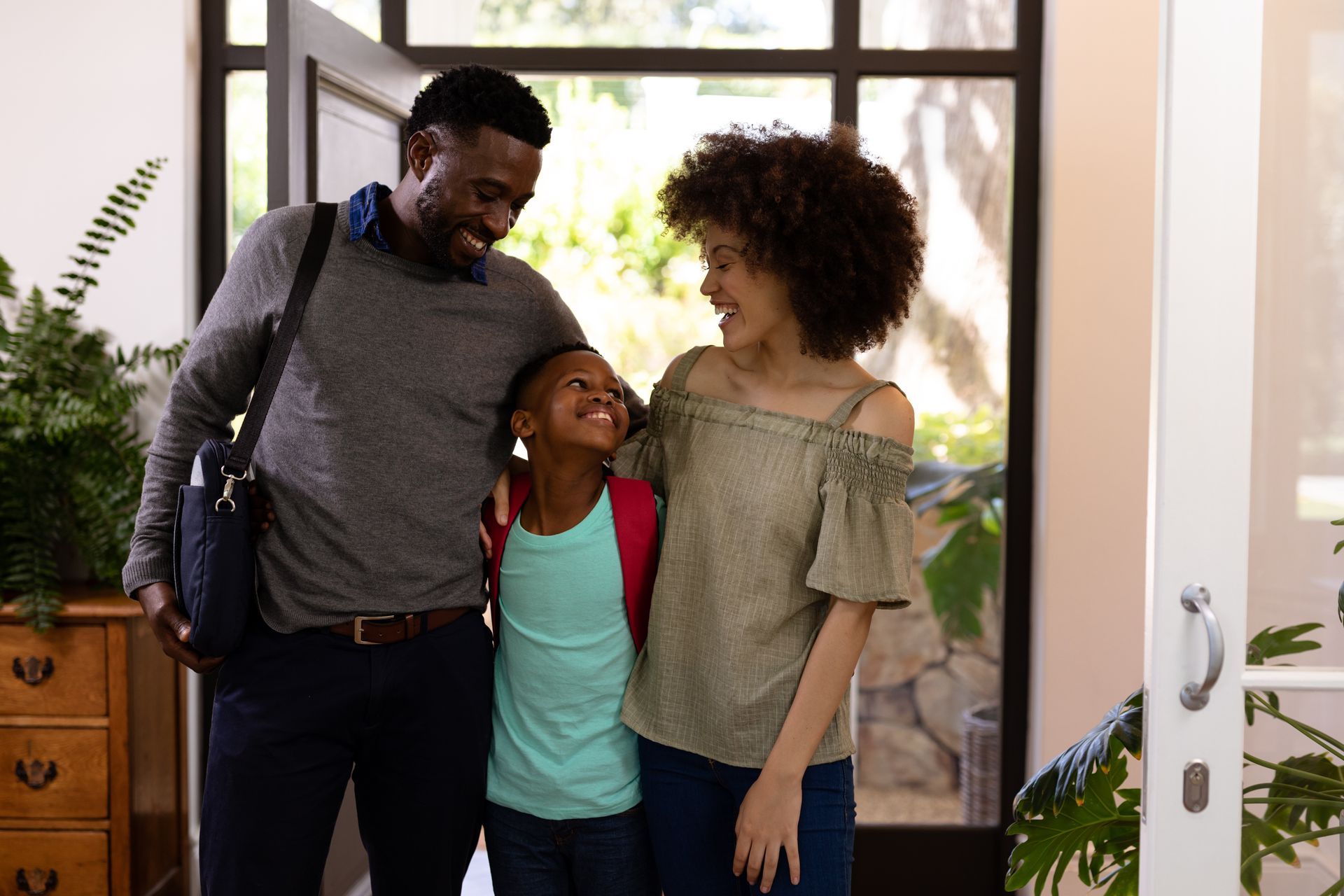 Family of three smiling, embracing near an open front door. Man has briefcase. Girl wears backpack.