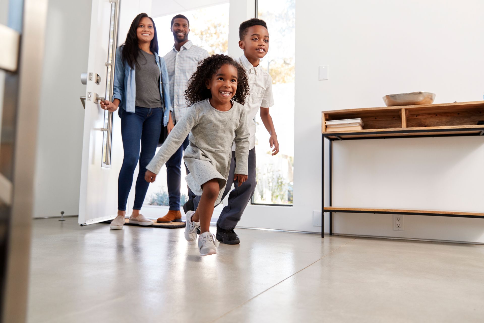 Family of four entering a bright home. Smiling children run forward; parents follow, smiling. White walls.
