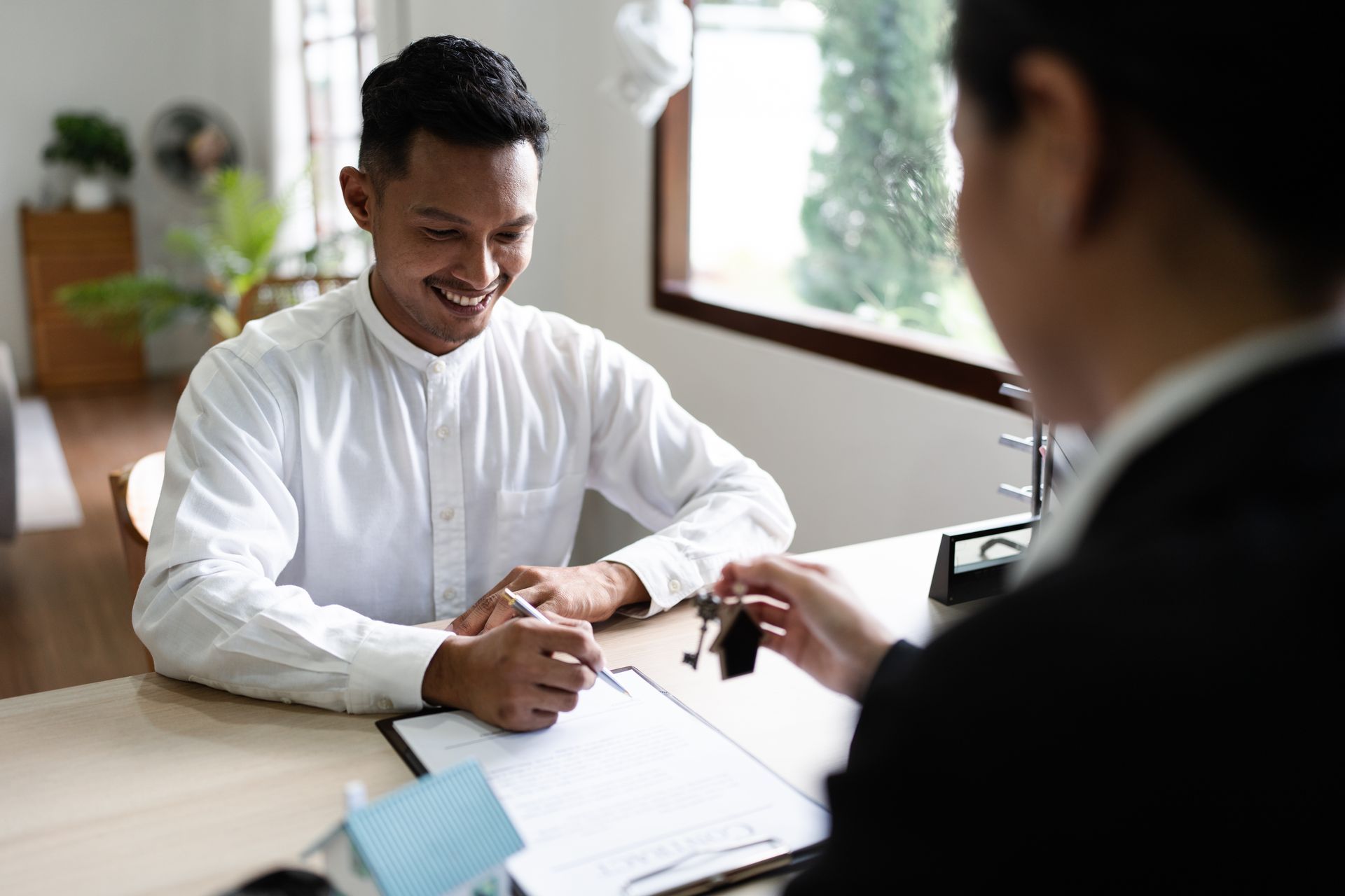 Man signs document as another person offers keys; indoor setting.
