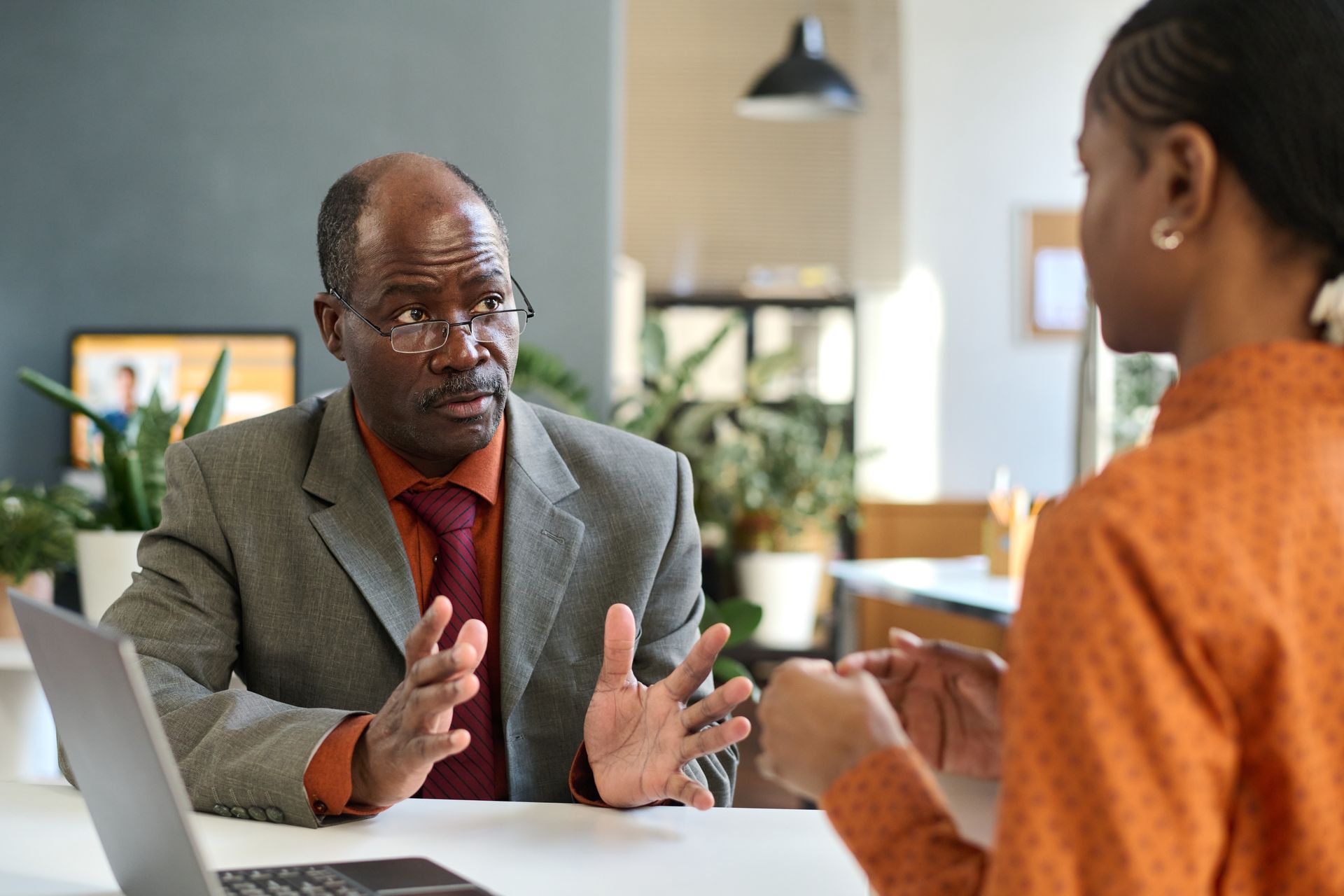 Man in suit gesturing while speaking to a person in an orange top. Office setting with laptop.