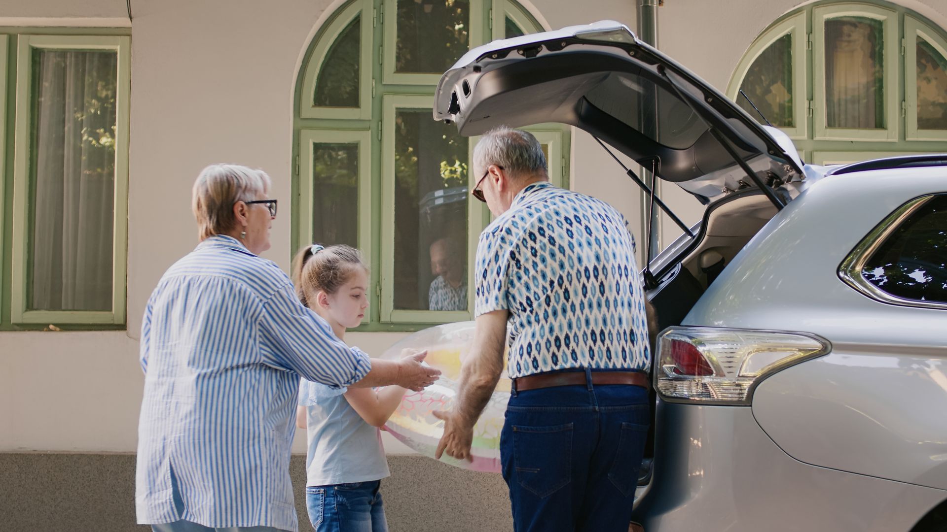 Grandparents and child loading items into a car trunk outside a building.
