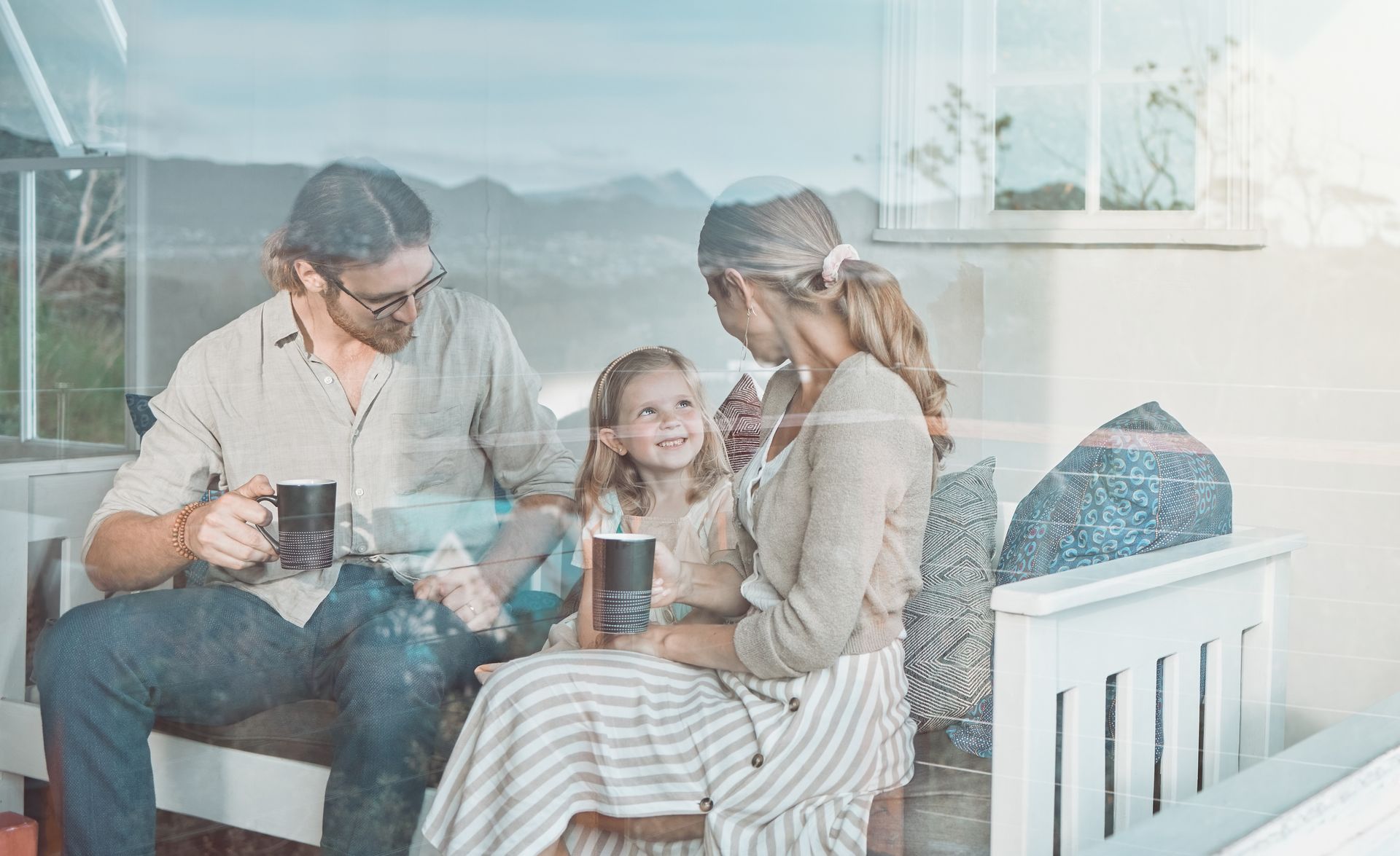Family sitting outdoors, smiling, holding mugs, overlooking a mountainous landscape.