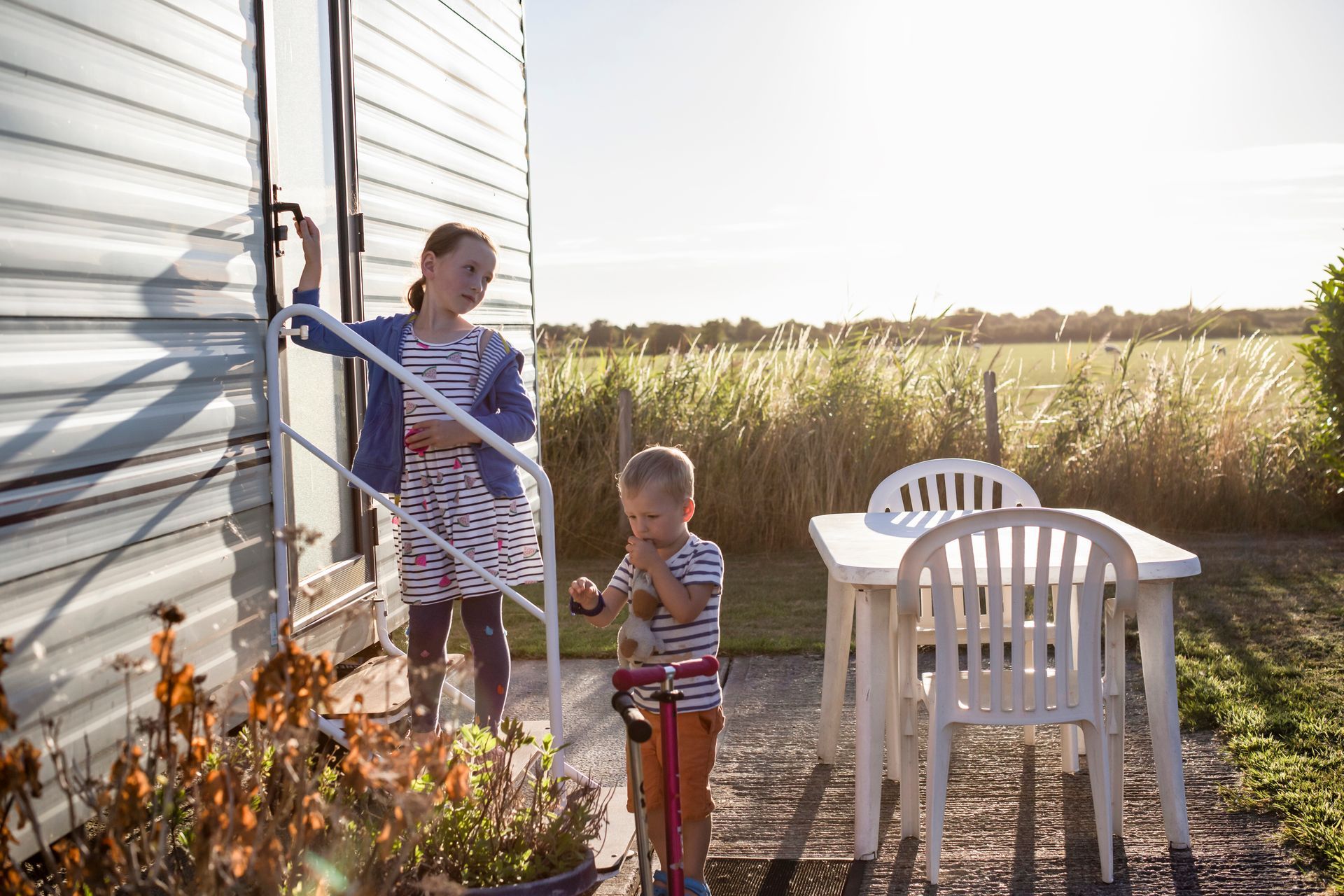 Two children near a trailer. One on steps, one with a scooter. Sunny outdoor setting.