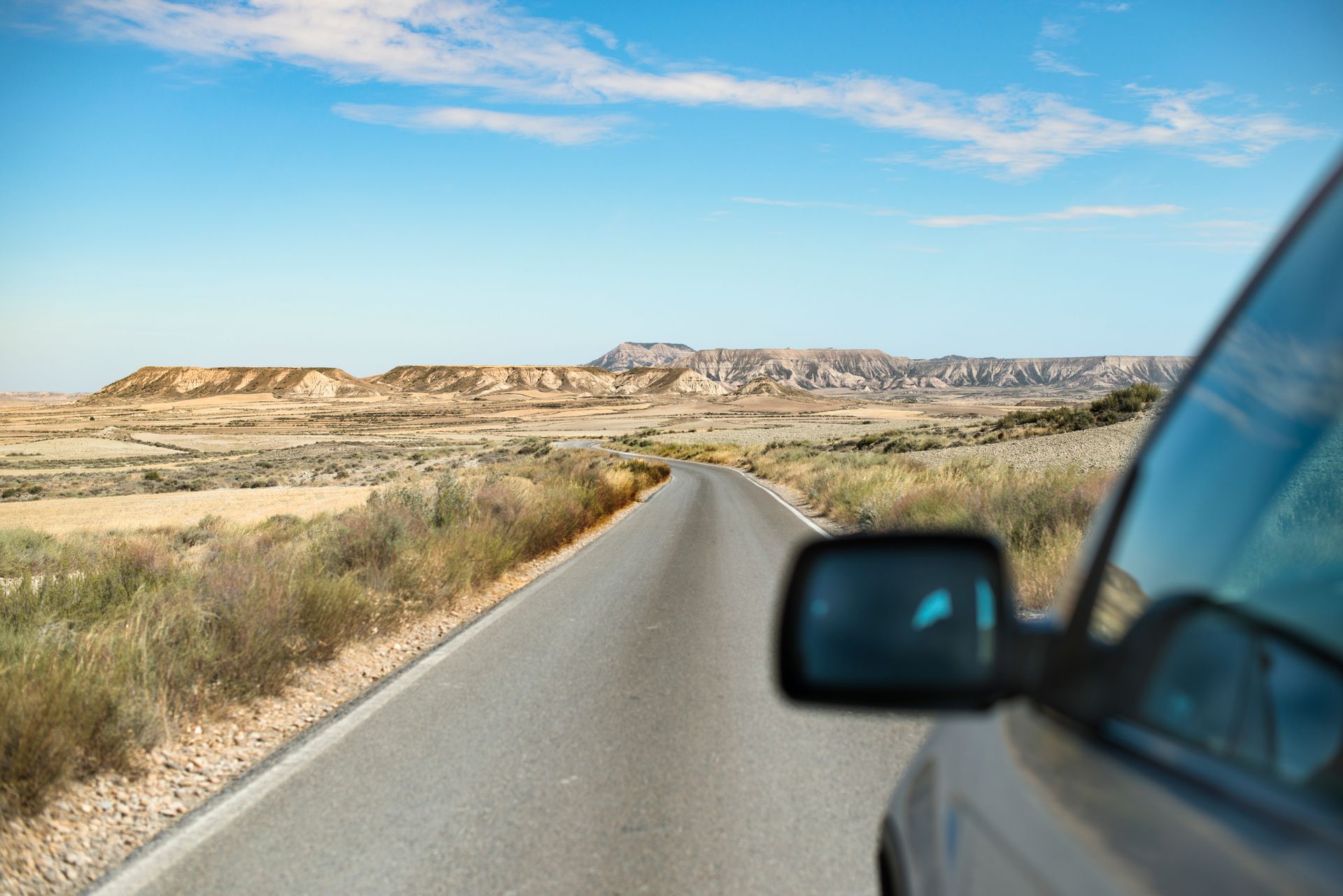 Road through a desert landscape, car side mirror in view, under a blue sky.