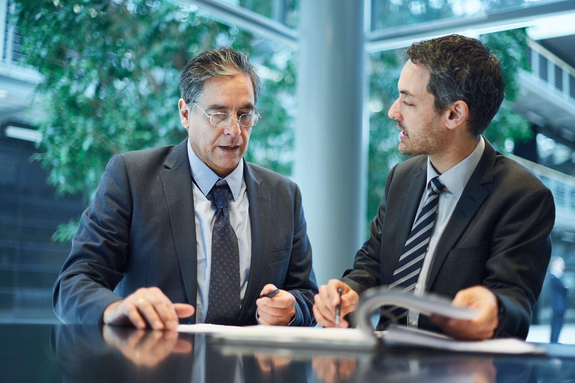 Two men in suits at a table, looking at documents and discussing. Modern office setting.