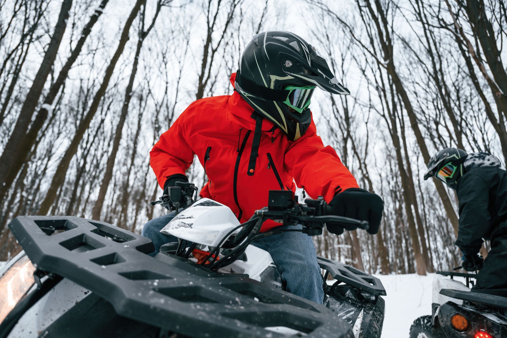 Person in red jacket on an ATV, driving in a snowy forest. Another ATV is nearby.