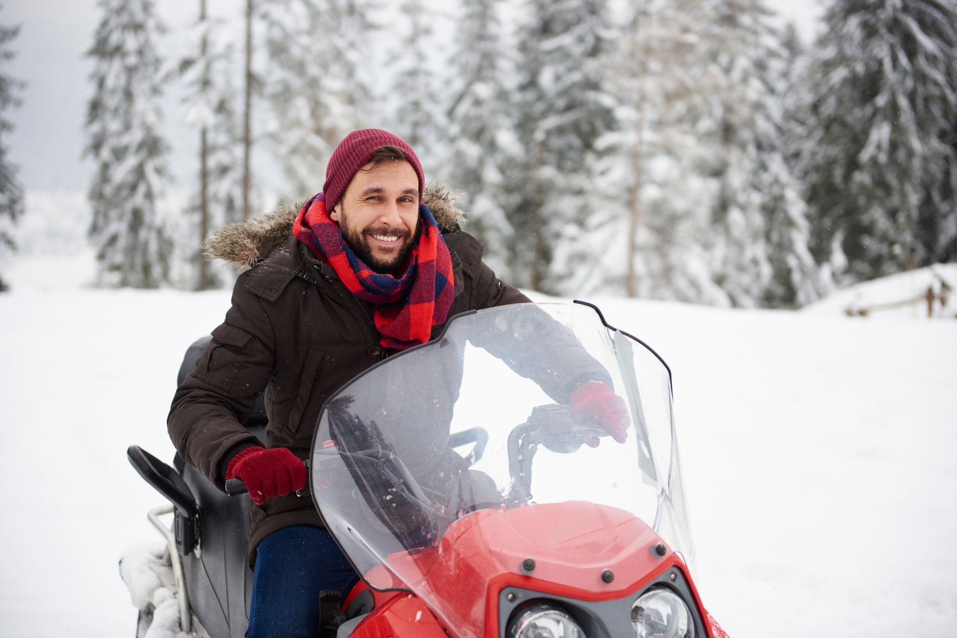 Man on a red snowmobile, smiling in a snowy forest.