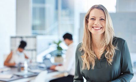 Blonde woman smiling at camera in office setting with coworkers blurred in background.